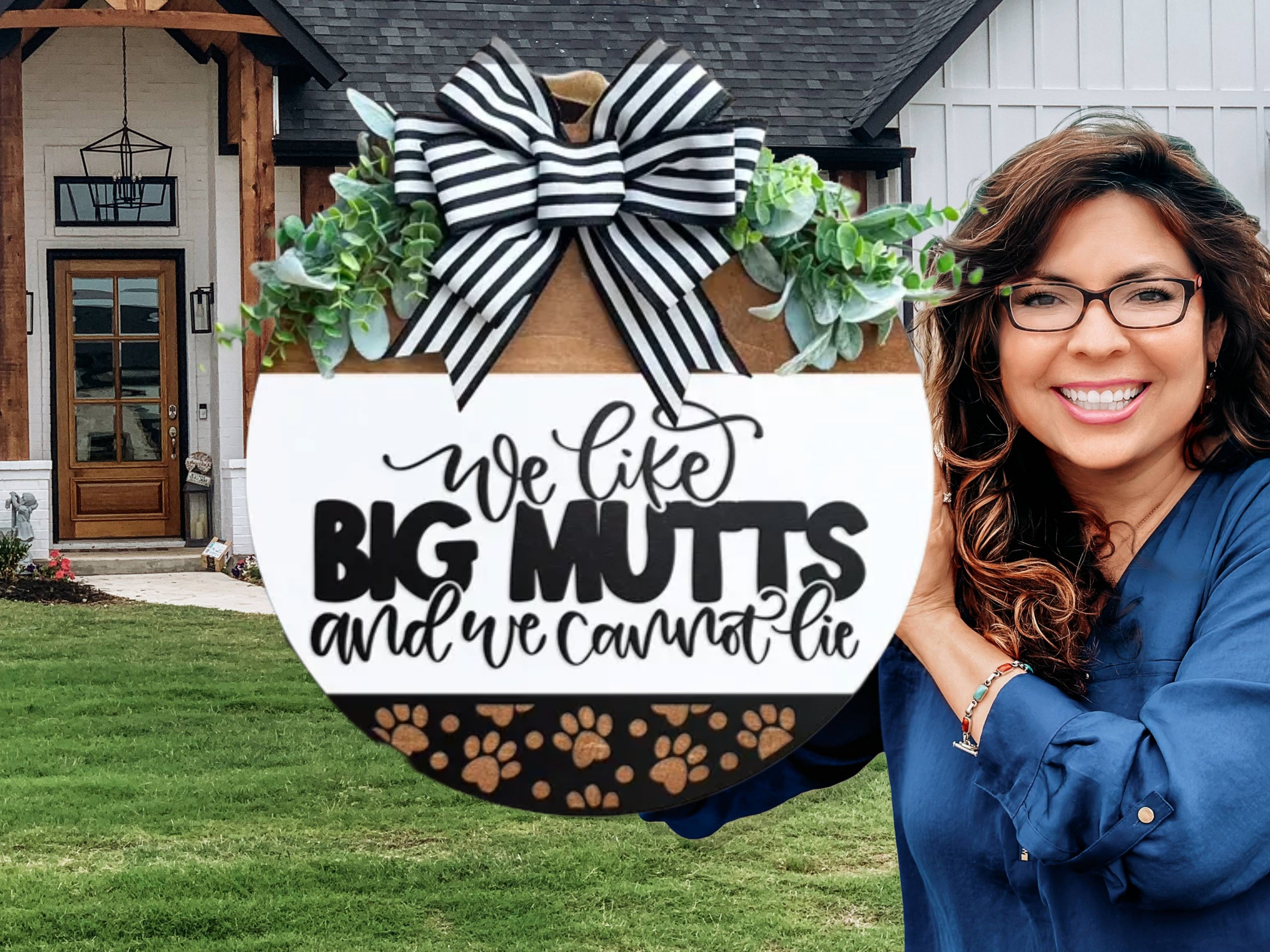 A smiling woman holds a round sign with a black-and-white bow and greenery that reads We like big mutts and we cannot lie. She stands in front of a modern house with a lawn.