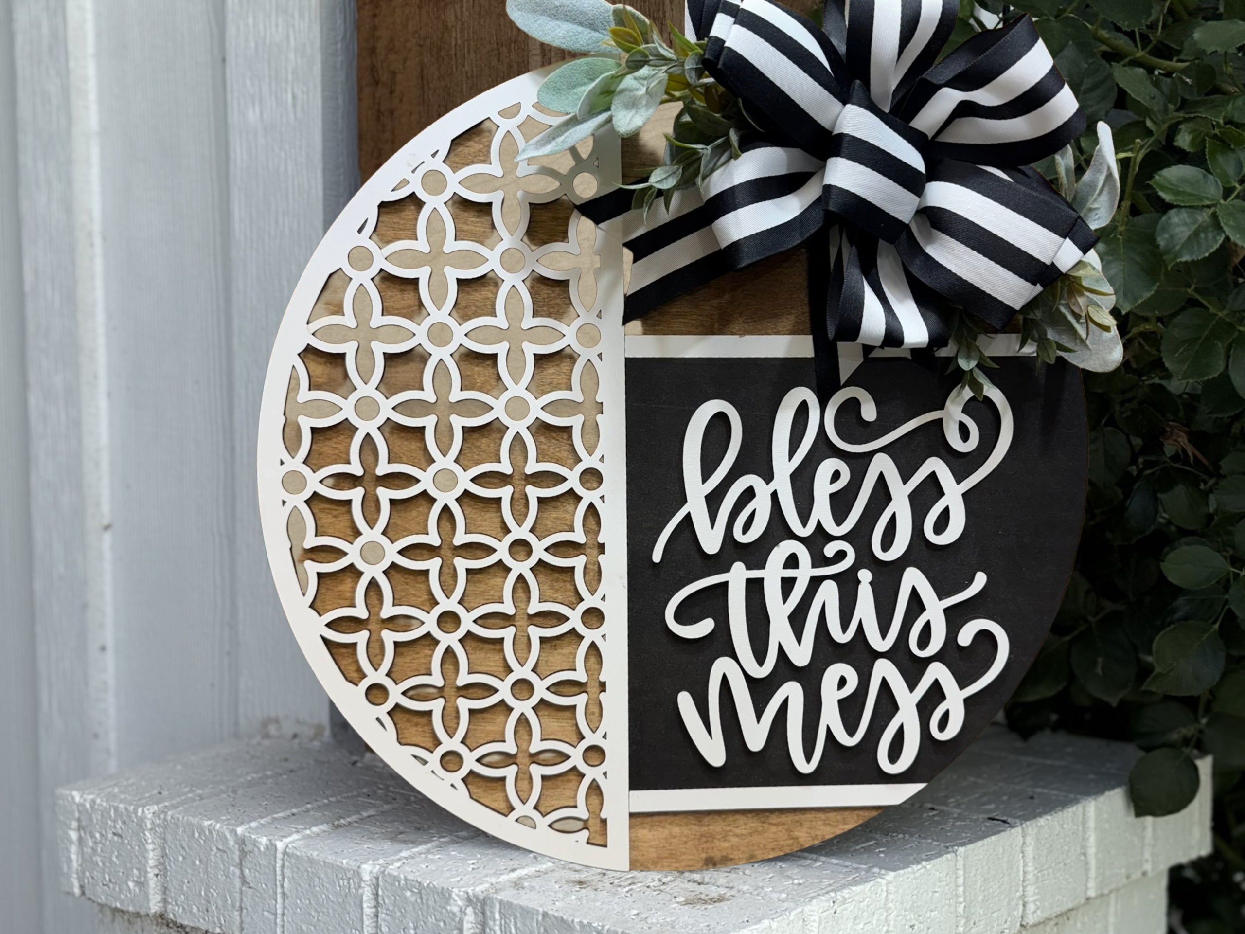 A round wooden sign with a white decorative lattice on the left and “bless this mess” in white script on black on the right, topped with greenery and a black-and-white striped bow, leans against a wall.