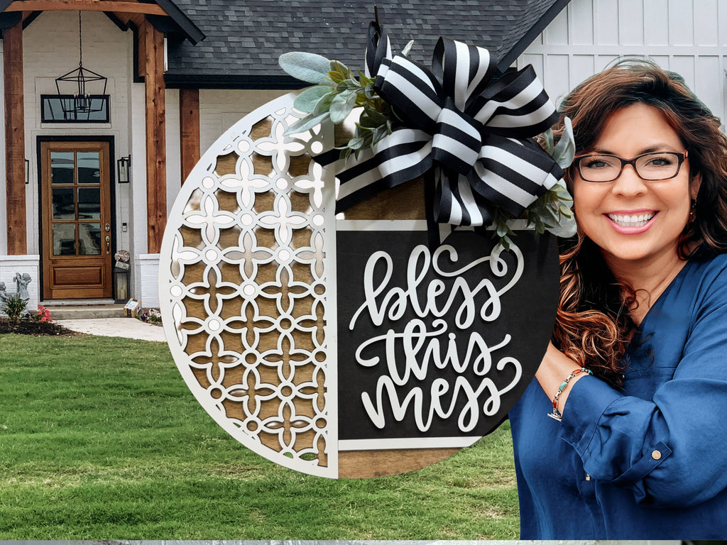A smiling woman with long dark hair and glasses holds a decorative round sign reading bless this mess in front of a modern farmhouse with white siding and wooden accents.