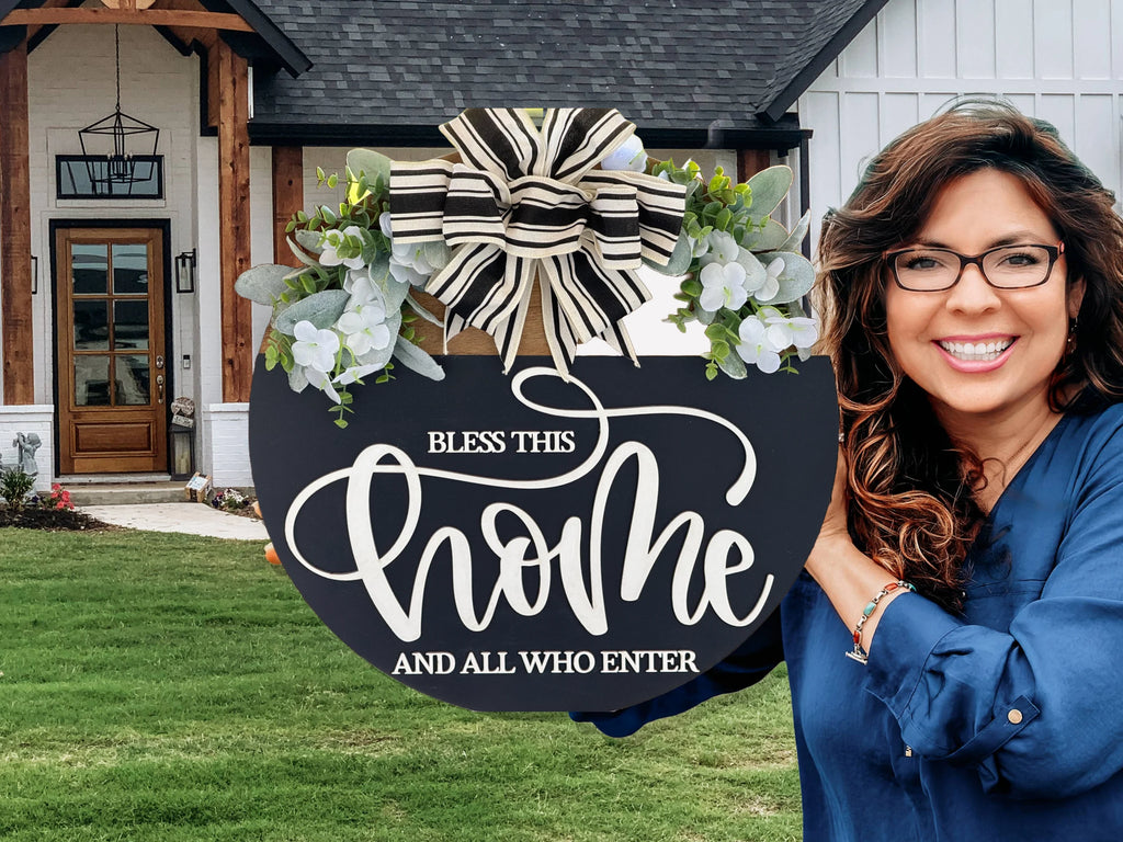 A smiling woman with wavy brown hair and glasses holds a black circular sign decorated with flowers and a black-and-white striped bow. The sign reads, Bless this home and all who enter. She stands in front of a modern house with a wooden door.
