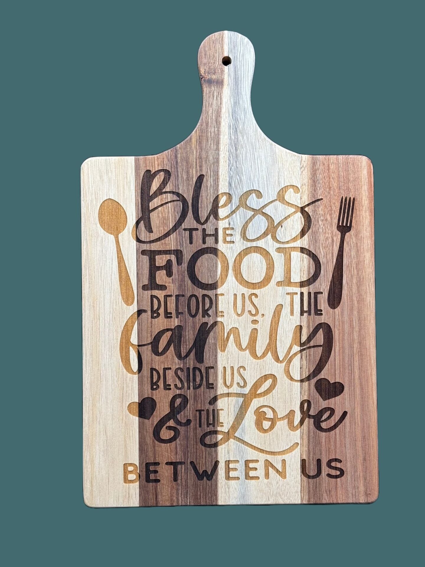 A wooden cutting board with vertical stripes in light and dark wood tones. It features an engraved decorative text: Bless the food before us, the family beside us & the love between us, along with spoon, fork, and heart symbols. The board has a handle with a hole for hanging.