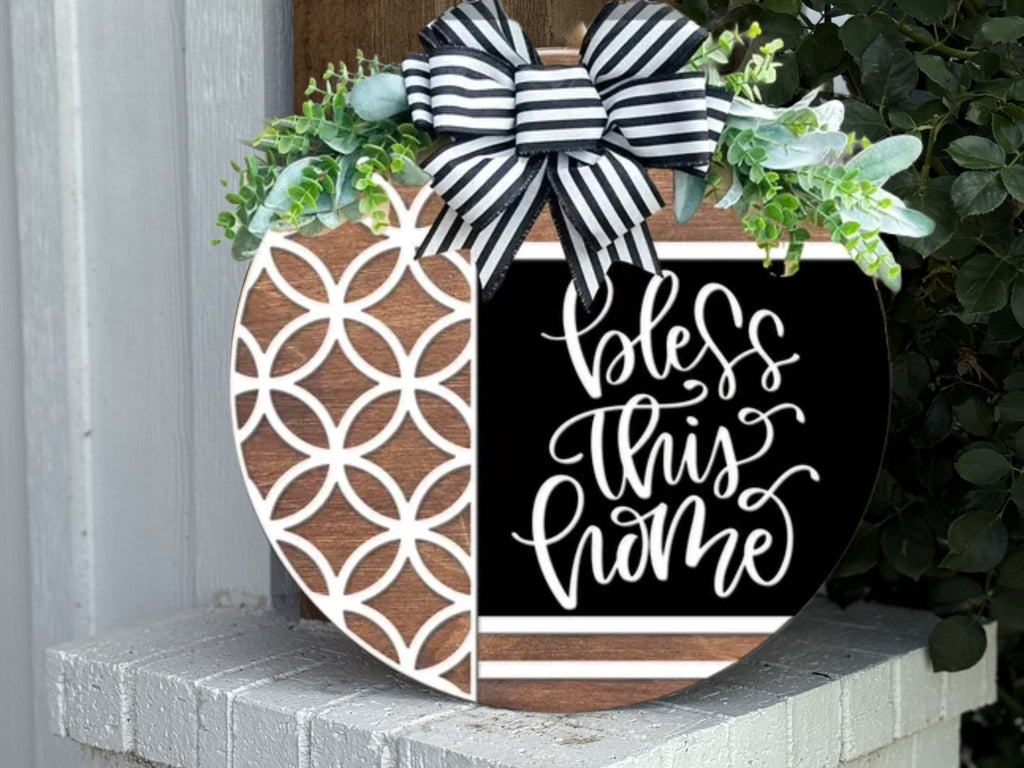 A round wooden sign decorated with a black and white striped bow, green leaves, a geometric pattern on the left, and the words “bless this home” in script on the black right side, sits on a white brick surface.