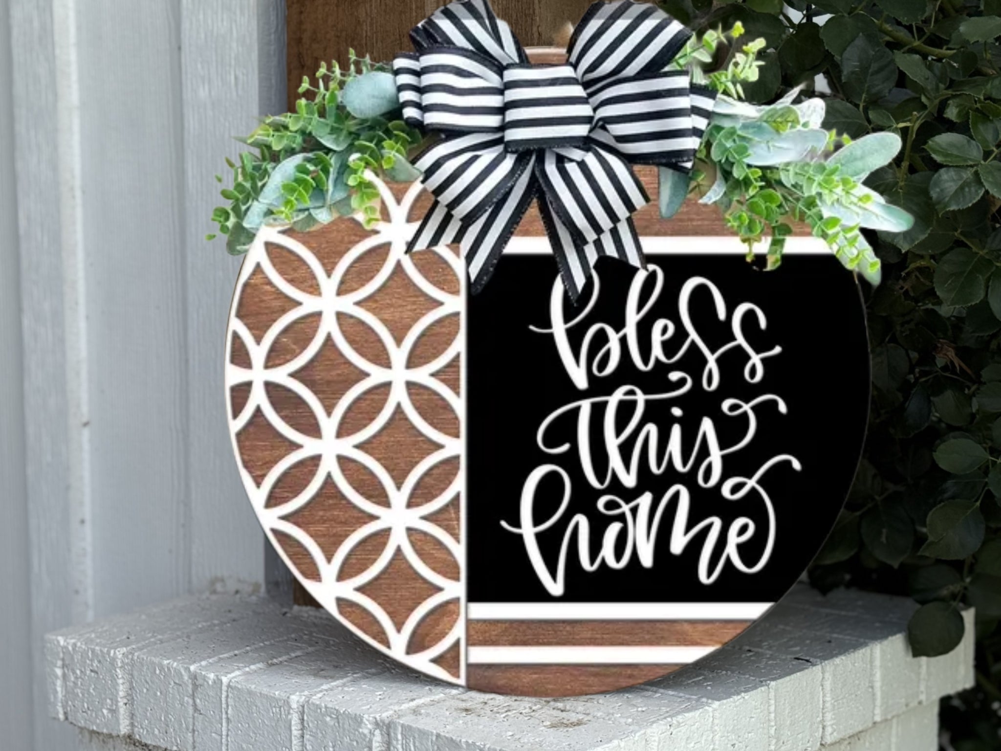 A round wooden sign decorated with a black and white striped bow, green leaves, a geometric pattern on the left, and the words “bless this home” in script on the black right side, sits on a white brick surface.