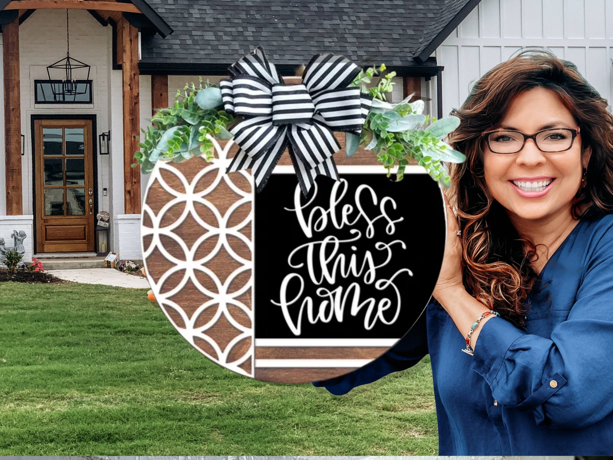 A smiling woman with long brown hair and glasses holds a round decorative sign that says bless this home in front of a modern house with a green lawn.