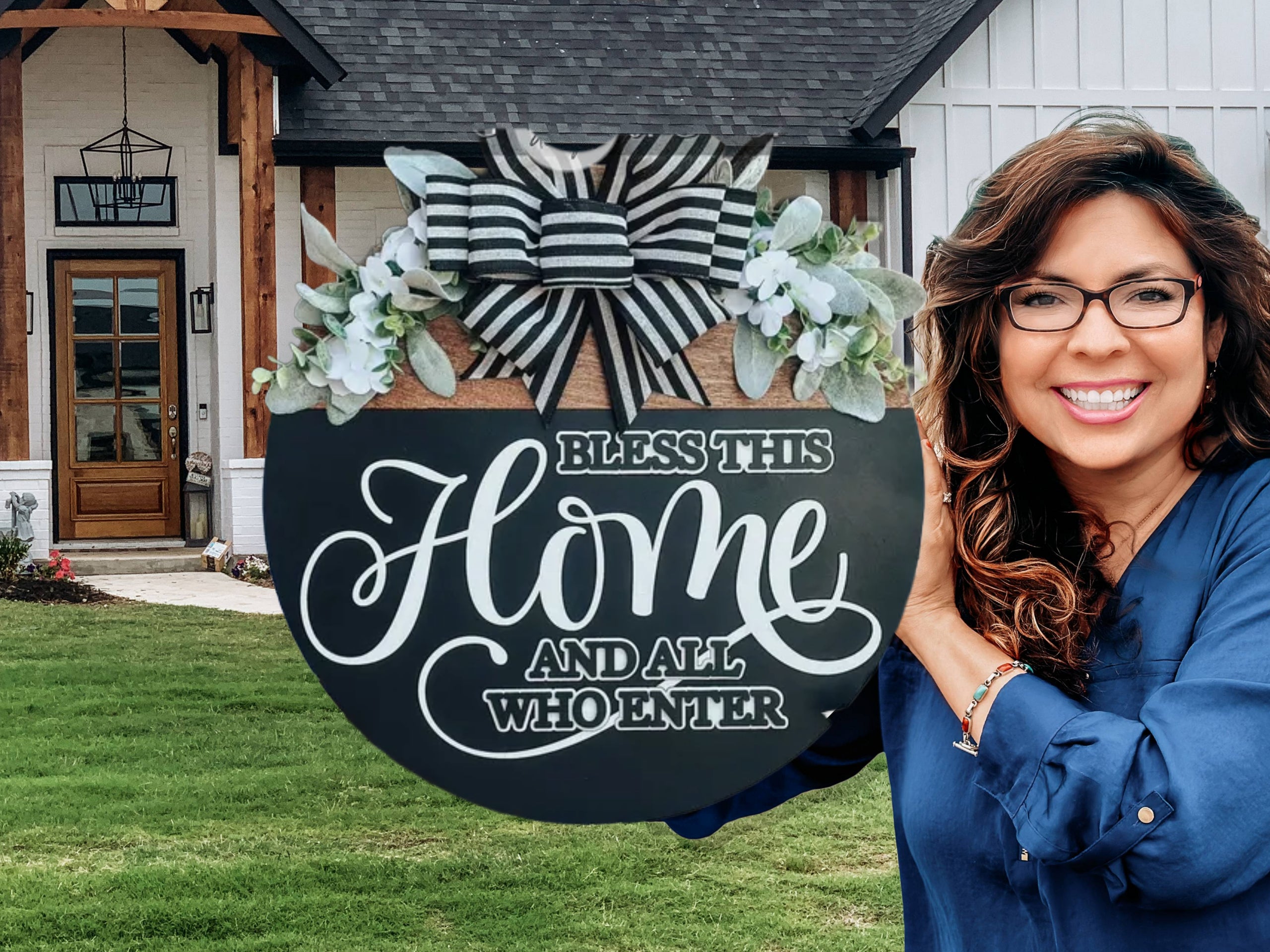 A smiling woman with long brown hair and glasses stands outside a house, holding a round door sign that reads, Bless this home and all who enter, decorated with greenery and a black-and-white striped bow.