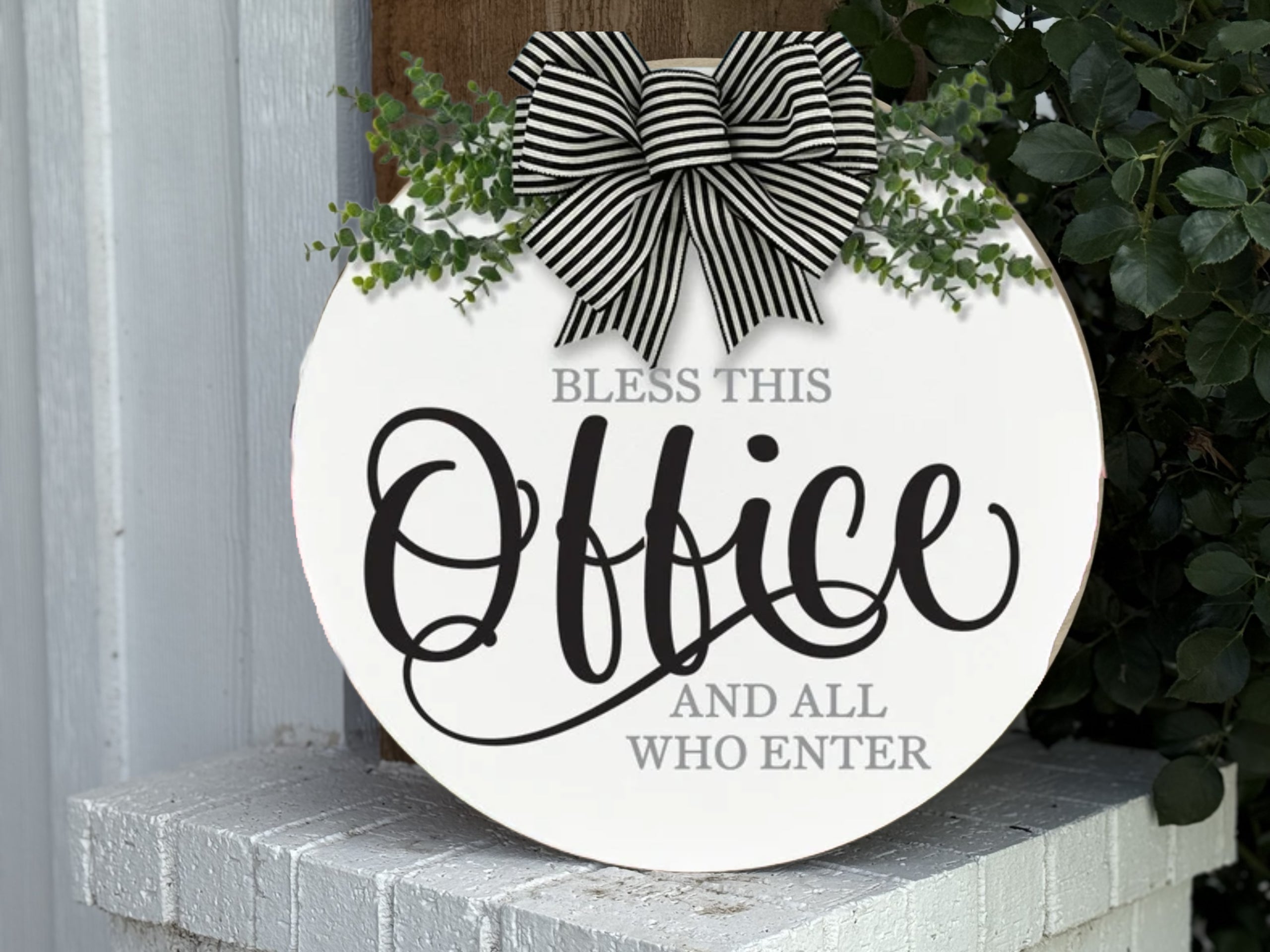 A round white sign with greenery and a black-and-white striped bow reads, Bless this office and all who enter in elegant black script. The sign sits on a white brick ledge next to a wooden post and leafy plant.