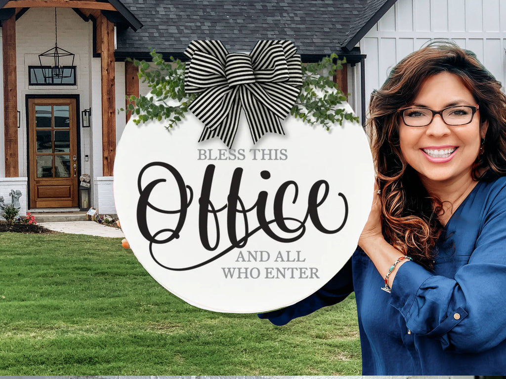 A smiling woman in glasses holds a round sign decorated with a striped bow and greenery that reads, “Bless this office and all who enter.” She stands outside a modern house with a wooden porch and white siding.