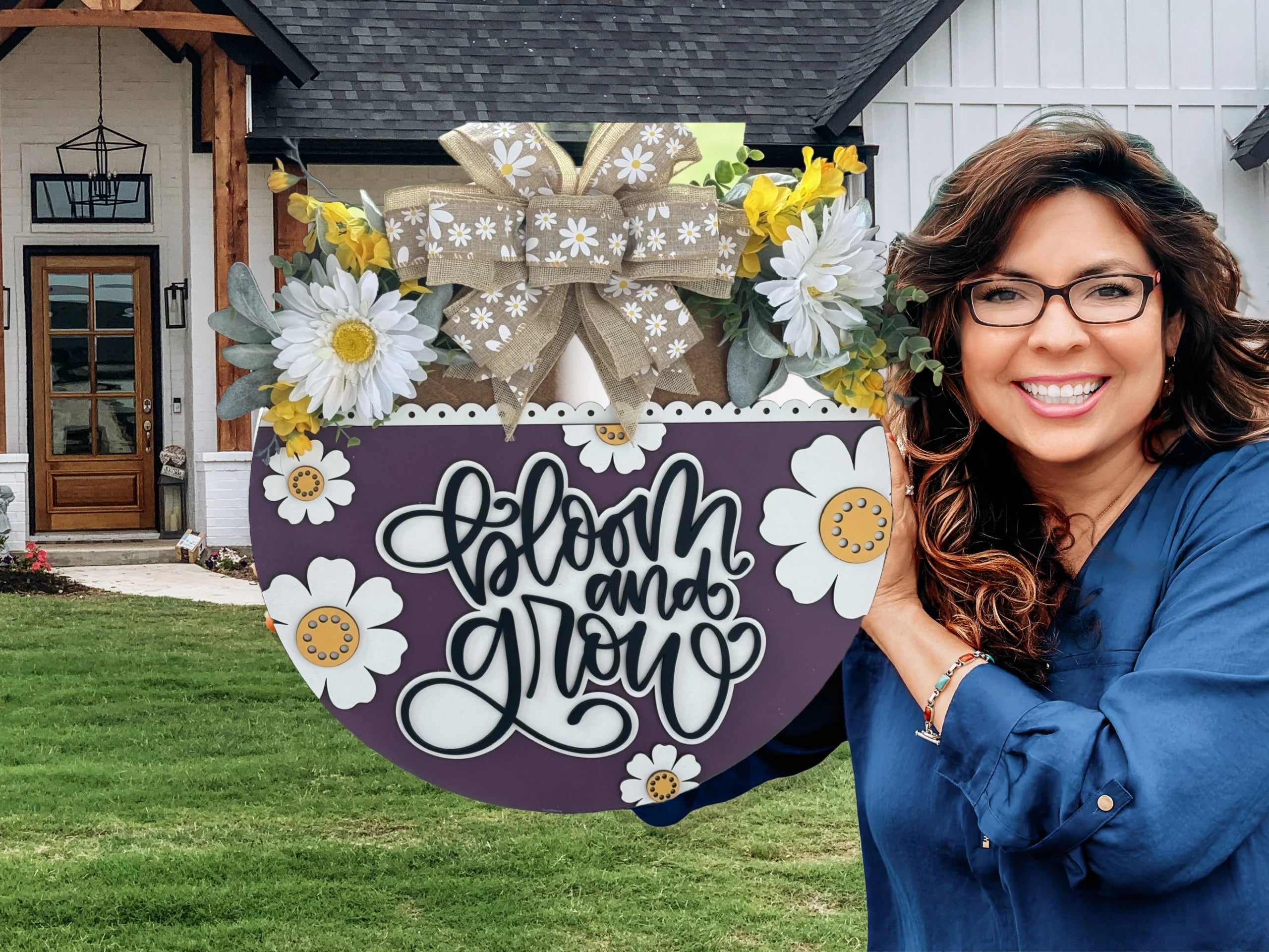 A woman with long, wavy hair and glasses smiles while holding a circular sign decorated with white daisies and a large beige bow. The sign reads Bloom and Grow in cursive. She stands in front of a modern house with a wooden door and large windows, surrounded by green grass.