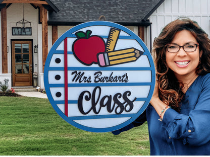 A smiling woman with wavy brown hair holds a round sign that says Mrs. Burkarts Class with drawings of an apple, pencil, and notebook paper. She stands in front of a house with a neatly kept yard.
