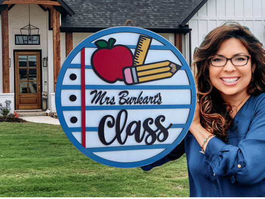 A smiling woman with wavy brown hair holds a round sign that says Mrs. Burkarts Class with drawings of an apple, pencil, and notebook paper. She stands in front of a house with a neatly kept yard.
