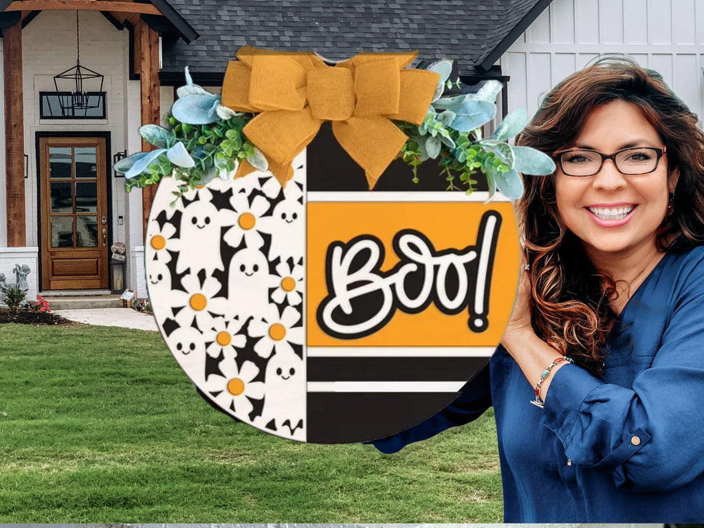A smiling woman with wavy brown hair and glasses holds a round Halloween sign that says Boo! in front of a modern house. The sign has cartoon ghosts, black stripes, and a yellow bow with greenery on top.