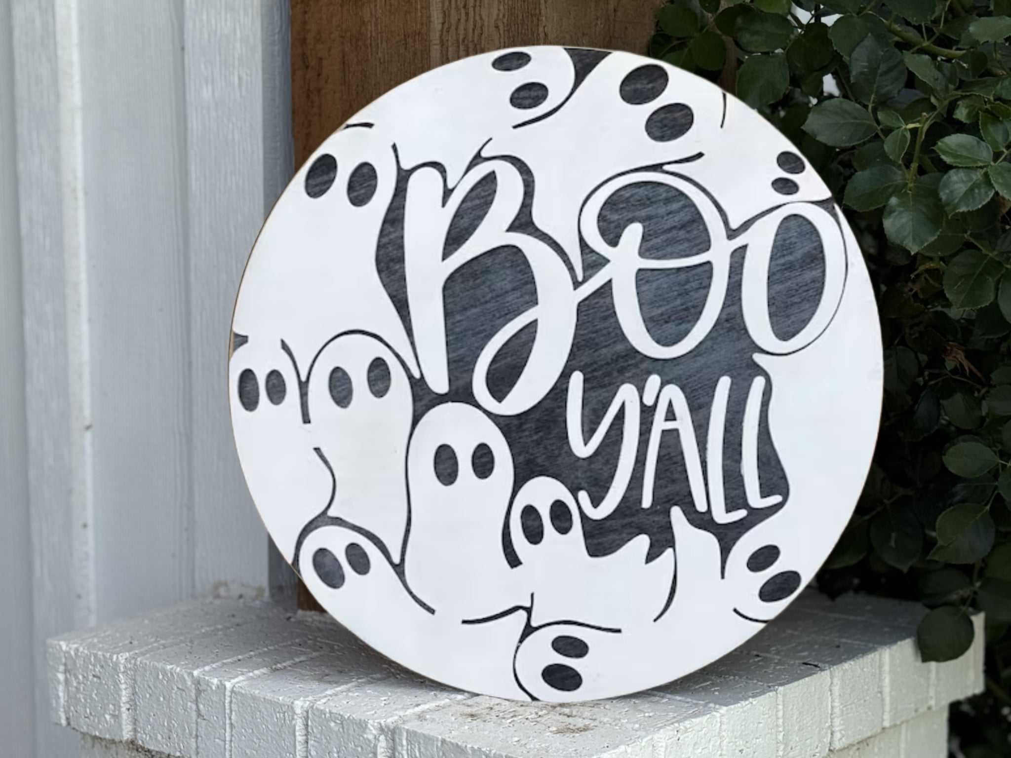 A round sign with the words Boo Yall in black, surrounded by hand-drawn cartoon ghosts. The sign leans against a wooden post and brick ledge, with green leaves on the right.