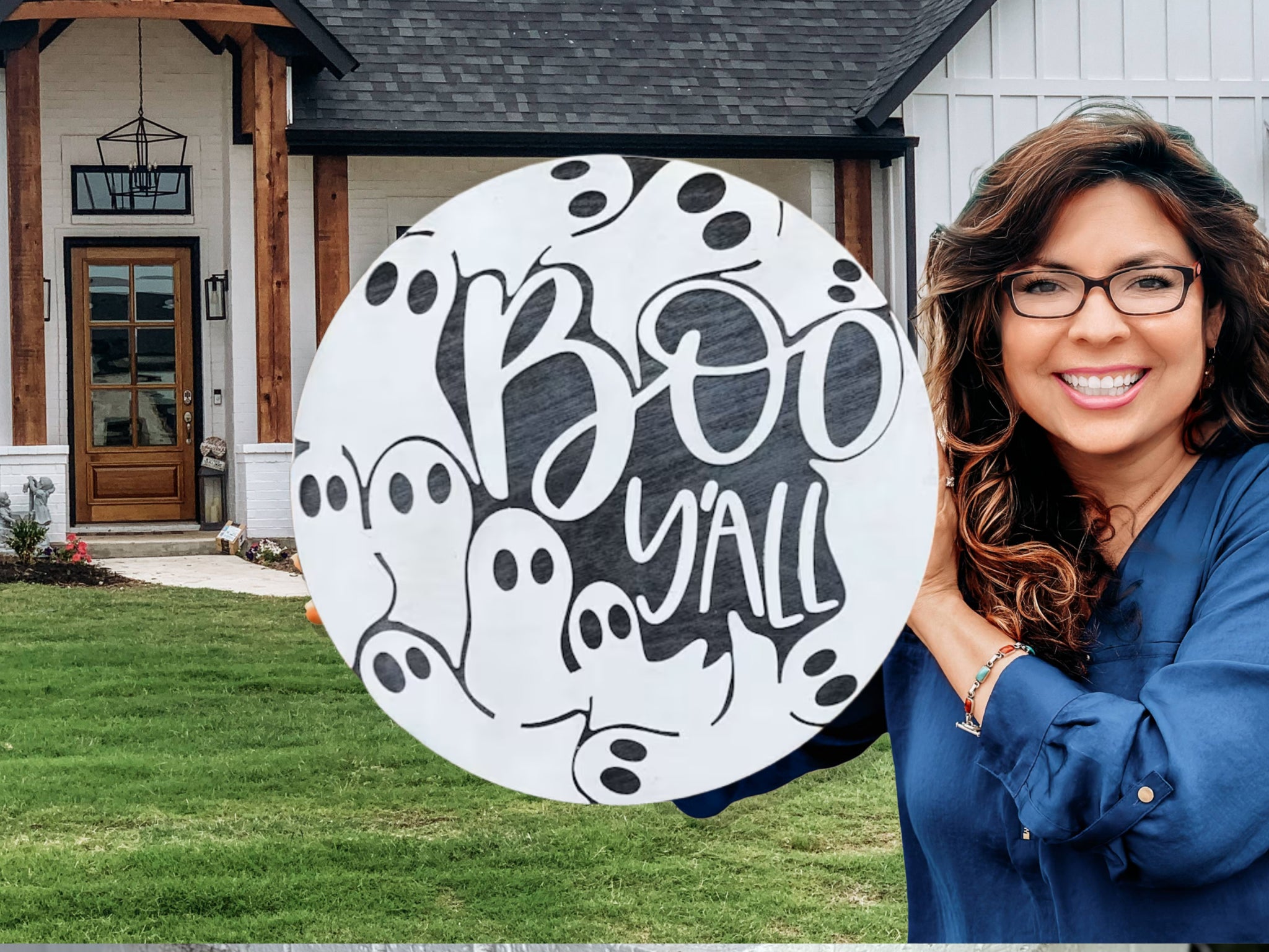 A smiling woman with glasses holds a round sign that says Boo Yall with black and white cartoon ghosts on it, standing in front of a house with green grass and wooden accents.