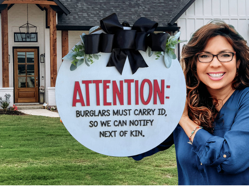 A smiling woman holds a round sign that reads, ATTENTION: BURGLARS MUST CARRY ID, SO WE CAN NOTIFY NEXT OF KIN. She stands in front of a modern house with a green lawn.