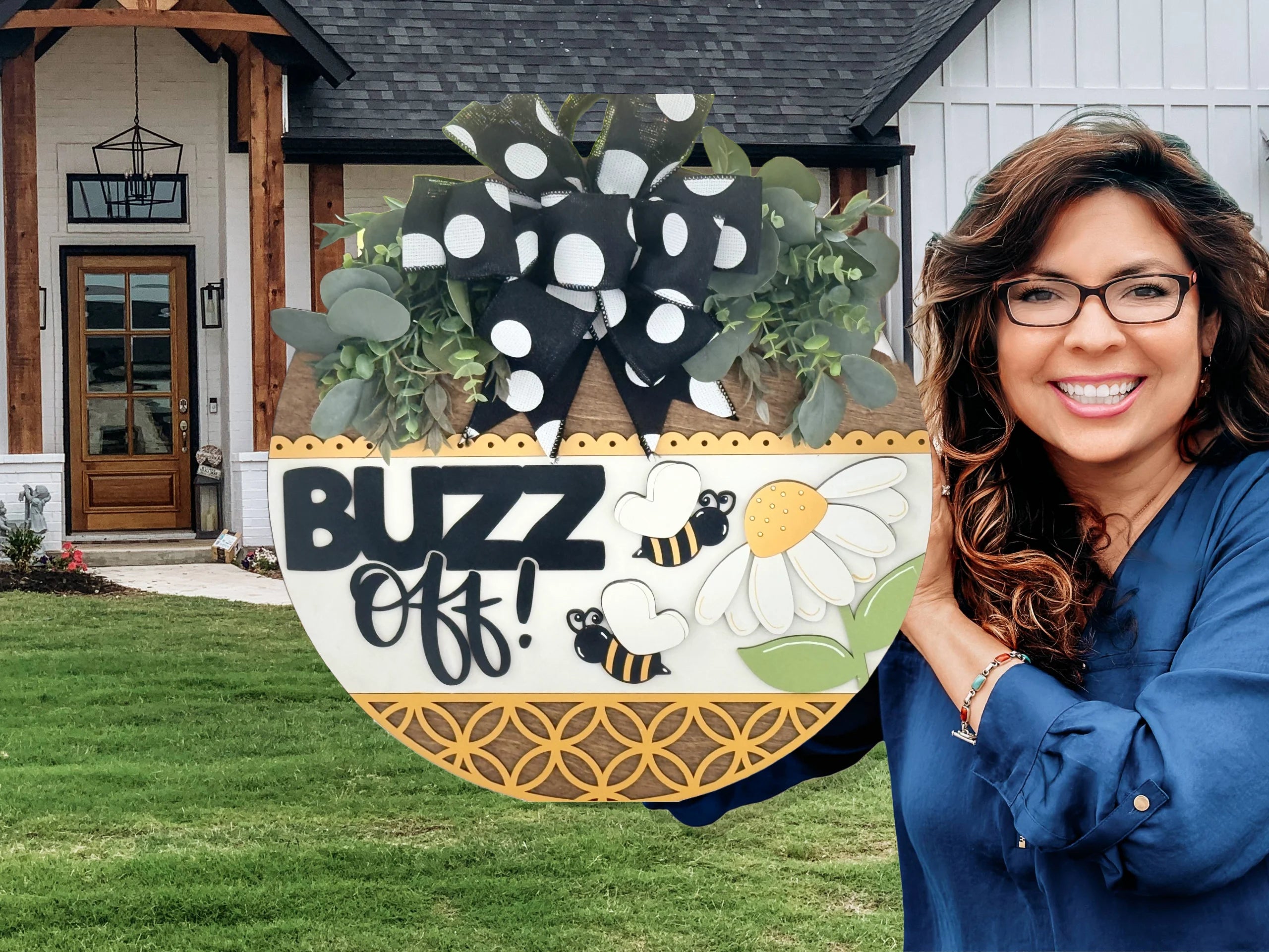 A smiling woman with wavy brown hair and glasses holds a round decorative sign with a daisy, two bees, greenery, and a large black-and-white polka dot bow. The sign says BUZZ off! She stands outside in front of a modern farmhouse.