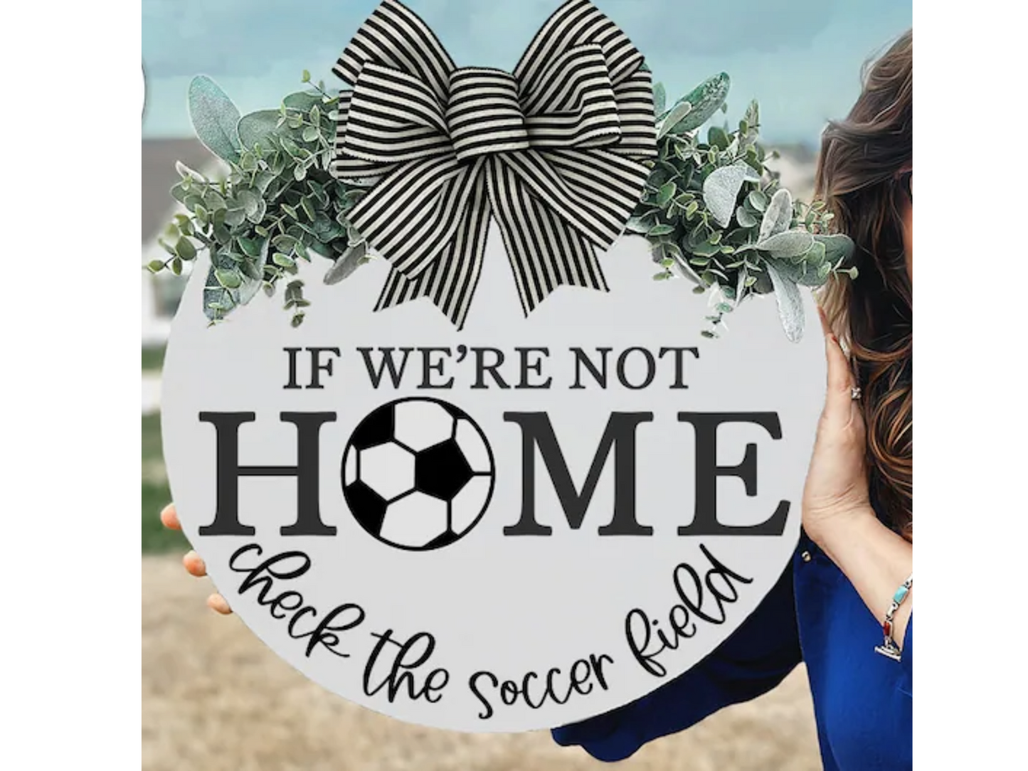 A person holds a round sign decorated with greenery and a black-and-white striped bow. The sign reads, If were not home, check the soccer field, with a soccer ball replacing the O in HOME.