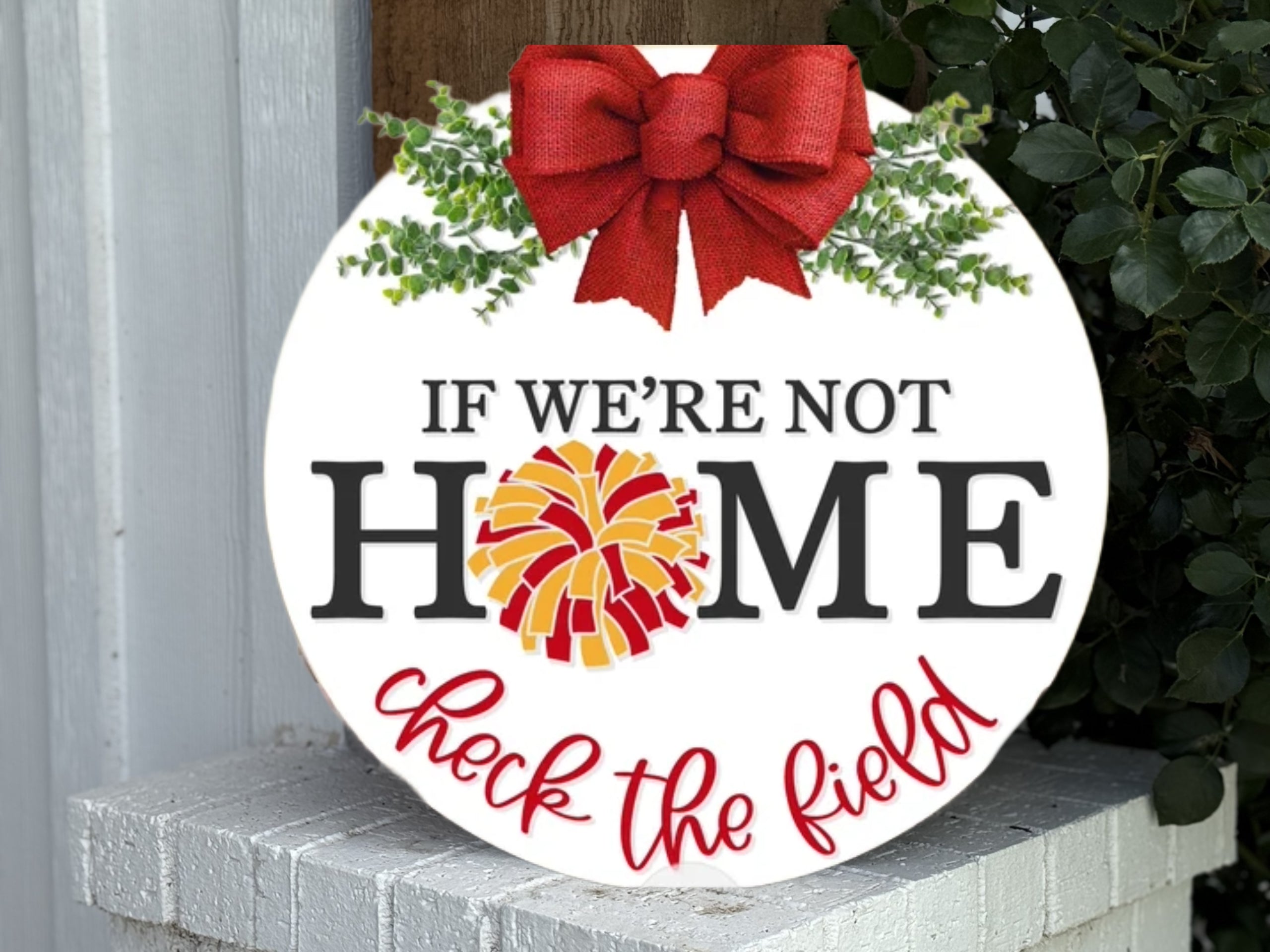 A round sign with a red bow and greenery on top reads, “If we’re not home” with “home” in black letters, the O replaced by a yellow and red pompom, and “check the field” written in red script below.