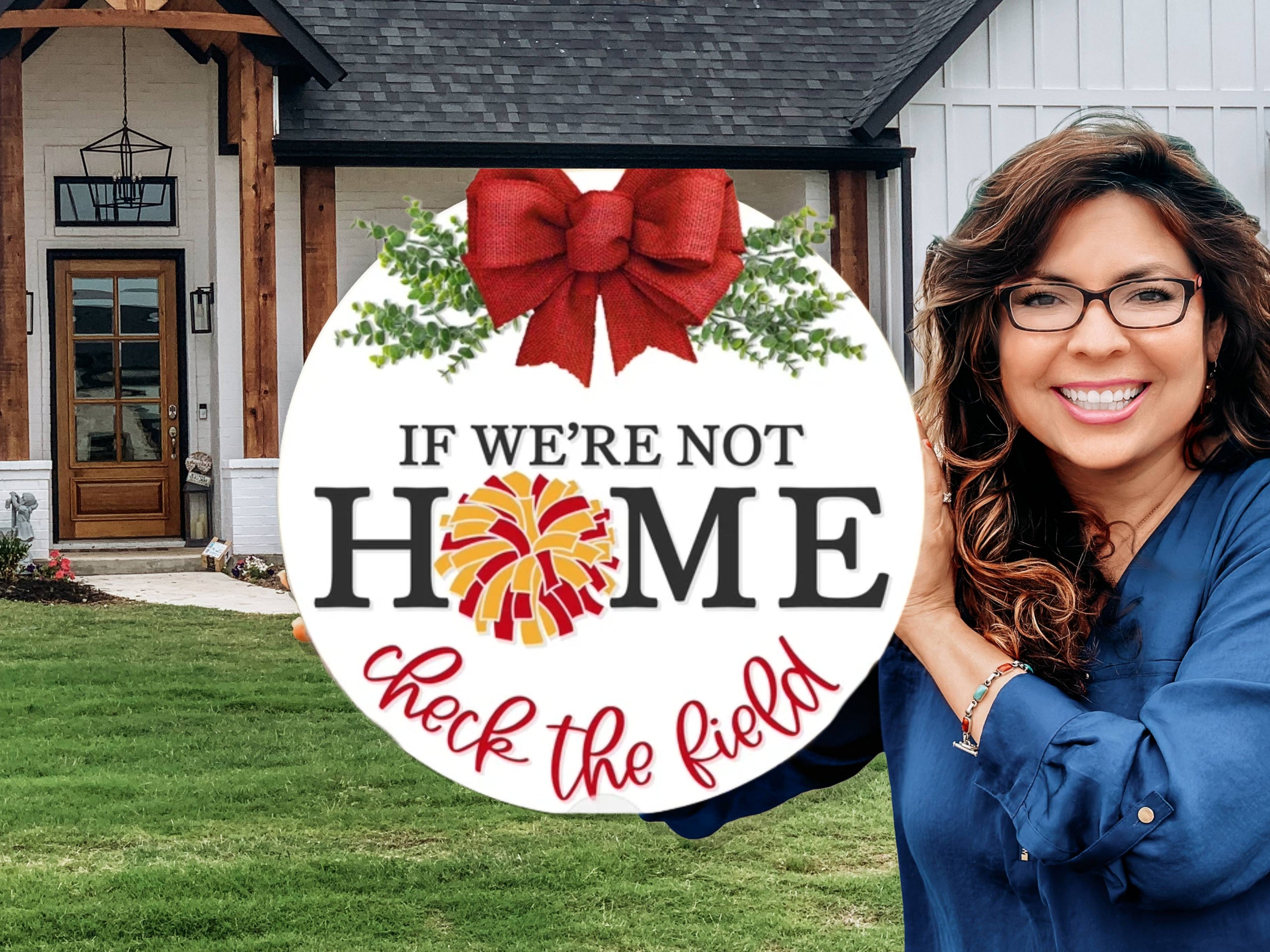 A smiling woman stands in front of a house, holding a round sign with a red bow that reads: If we’re not home, check the field with home decorated with a yellow and red pom-pom for the letter O.