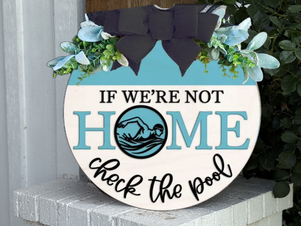 A round sign with flowers and a black bow reads: “IF WE’RE NOT HOME check the pool.” The word “HOME” features a swimming figure inside the letter O. The sign rests on a white brick surface near greenery.