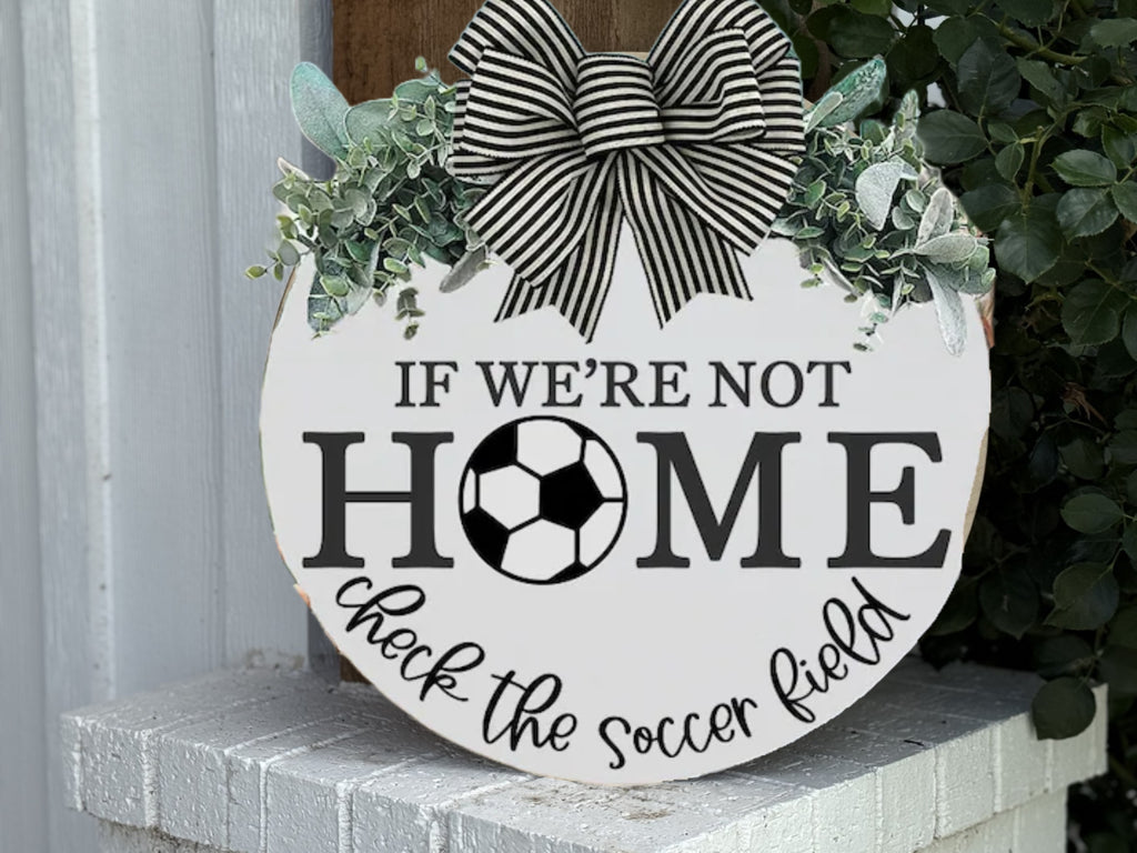 A round sign with a black-and-white striped bow and greenery reads: IF WERE NOT HOME check the soccer field, with a soccer ball illustration in place of the letter O in HOME.