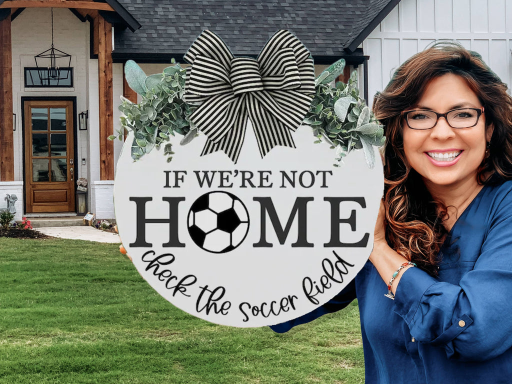 A smiling woman with long brown hair and glasses stands outside a house, holding a round sign with a bow that reads, IF WE’RE NOT HOME check the soccer field, with a soccer ball graphic in the word HOME.