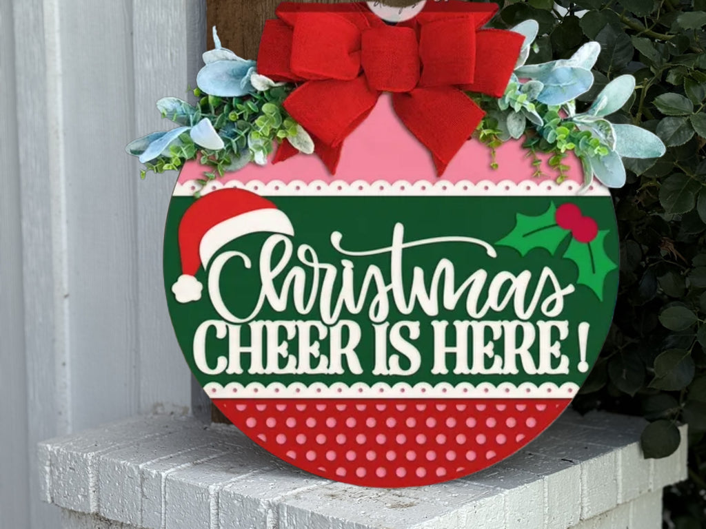 A festive round sign decorated with a red bow, greenery, and berries reads Christmas Cheer Is Here! in white letters, with a Santa hat and holly, resting on a white brick ledge.