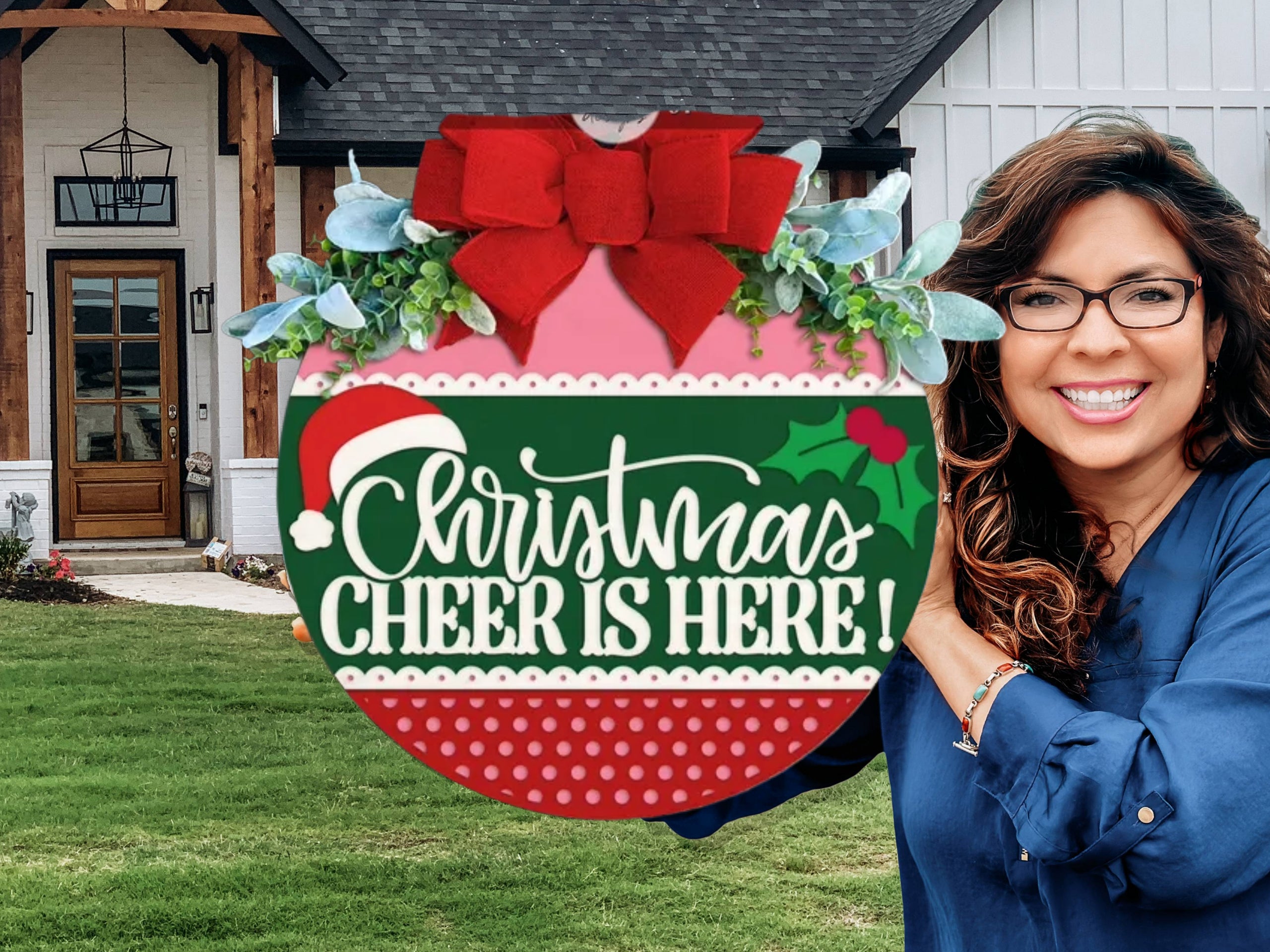 A smiling woman in glasses holds a large festive sign shaped like a Christmas ornament that says, Christmas Cheer is Here! in front of a modern house with a lawn.