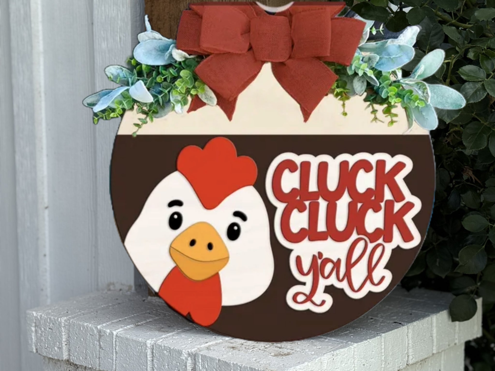 A round wooden sign with a red bow, greenery, and a cartoon chicken face. The sign reads CLUCK CLUCK y’all in bold red and white letters. It is displayed outdoors on a white brick surface.