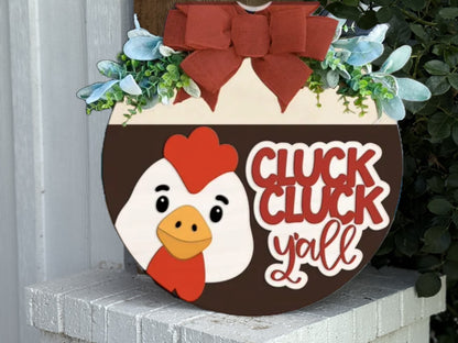 A round wooden sign with a red bow, greenery, and a cartoon chicken face. The sign reads CLUCK CLUCK y’all in bold red and white letters. It is displayed outdoors on a white brick surface.