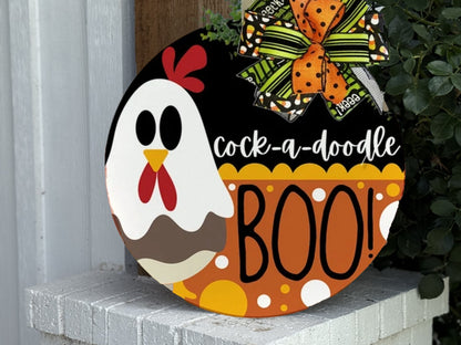 A round sign with a cartoon chicken, the text “cock-a-doodle BOO!”, orange polka dots, and a festive orange and green bow, displayed outdoors near a white wall and greenery.