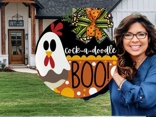 A round sign with a cartoon chicken, the text “cock-a-doodle BOO!”, orange polka dots, and a festive orange and green bow, displayed outdoors near a white wall and greenery.