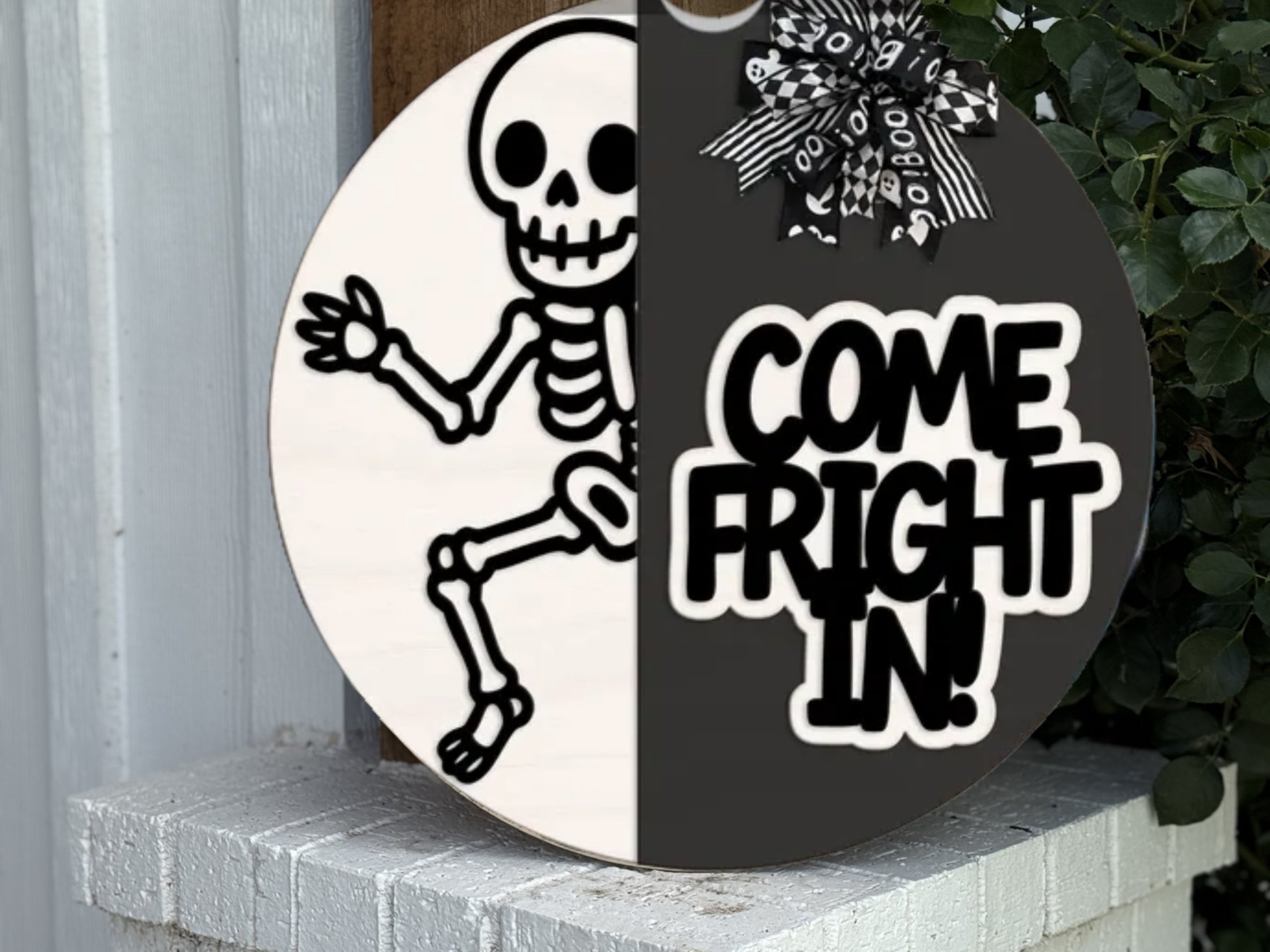 A round sign with a black-and-white split design: one side shows a cartoon skeleton, the other side says COME FRIGHT IN! in bold letters, topped with a black-and-white bow, leaning on a white brick step.