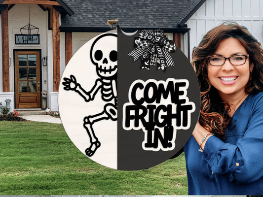 A smiling woman with wavy hair stands outside a modern house. In front of her is a round Halloween sign with a skeleton and the words Come Fright In! decorated with a black and white bow.