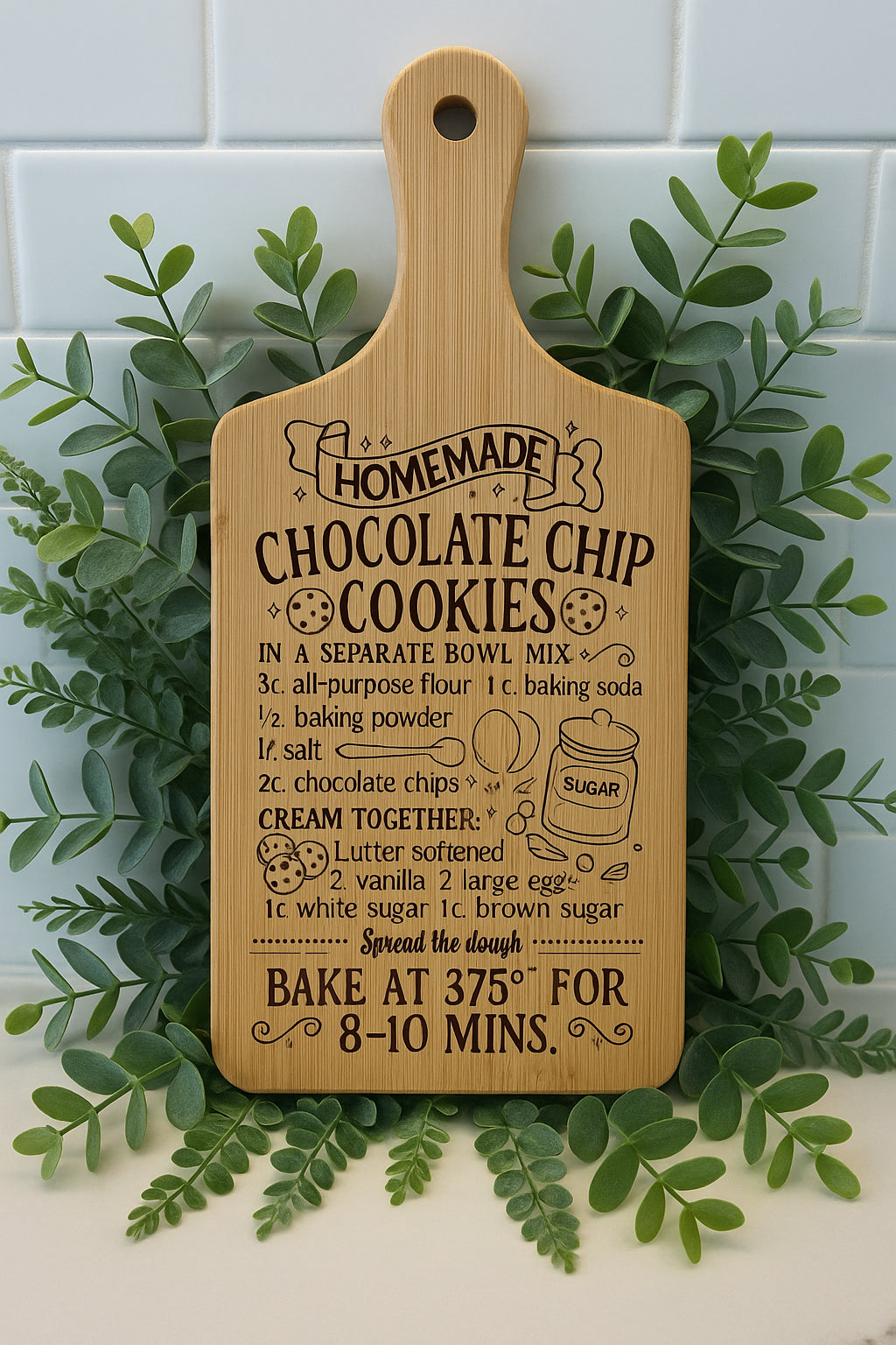 A wooden cutting board with a recipe for homemade chocolate chip cookies, surrounded by green leaves, is displayed against a white tiled background. The recipe includes ingredients and baking instructions.
