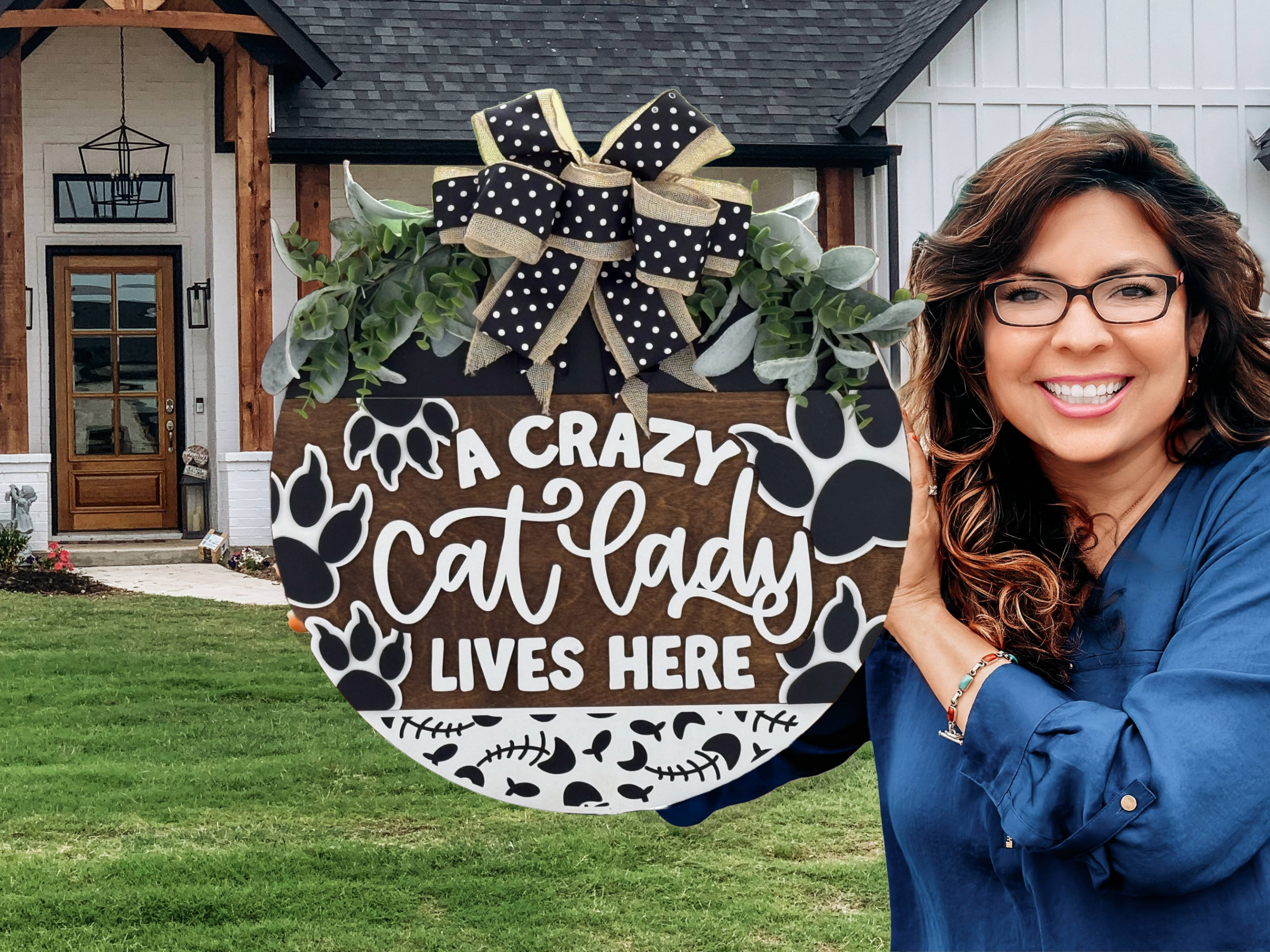 A smiling woman with wavy hair and glasses holds a round sign decorated with a bow, paw prints, and foliage that reads, A crazy cat lady lives here, standing outside a house with a wooden porch.