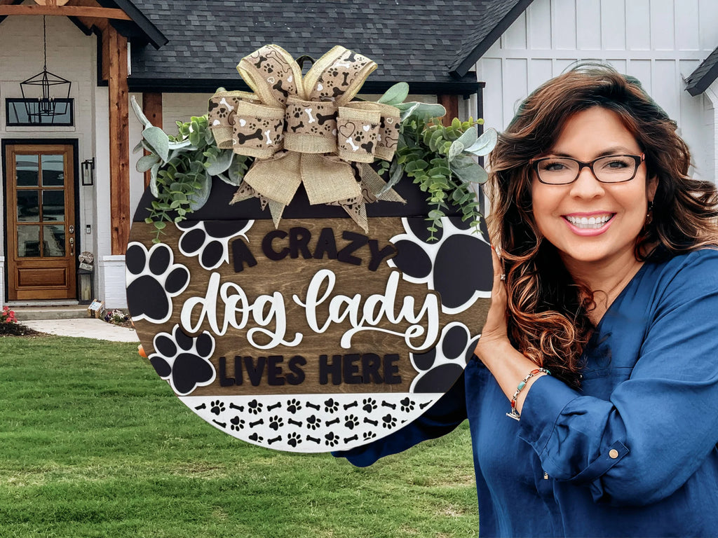 Smiling woman holding a round decorative sign that reads A Crazy Dog Lady Lives Here with paw prints and leaves. Shes standing in front of a modern house with a wooden door and green lawn. Shes wearing glasses and a blue top.