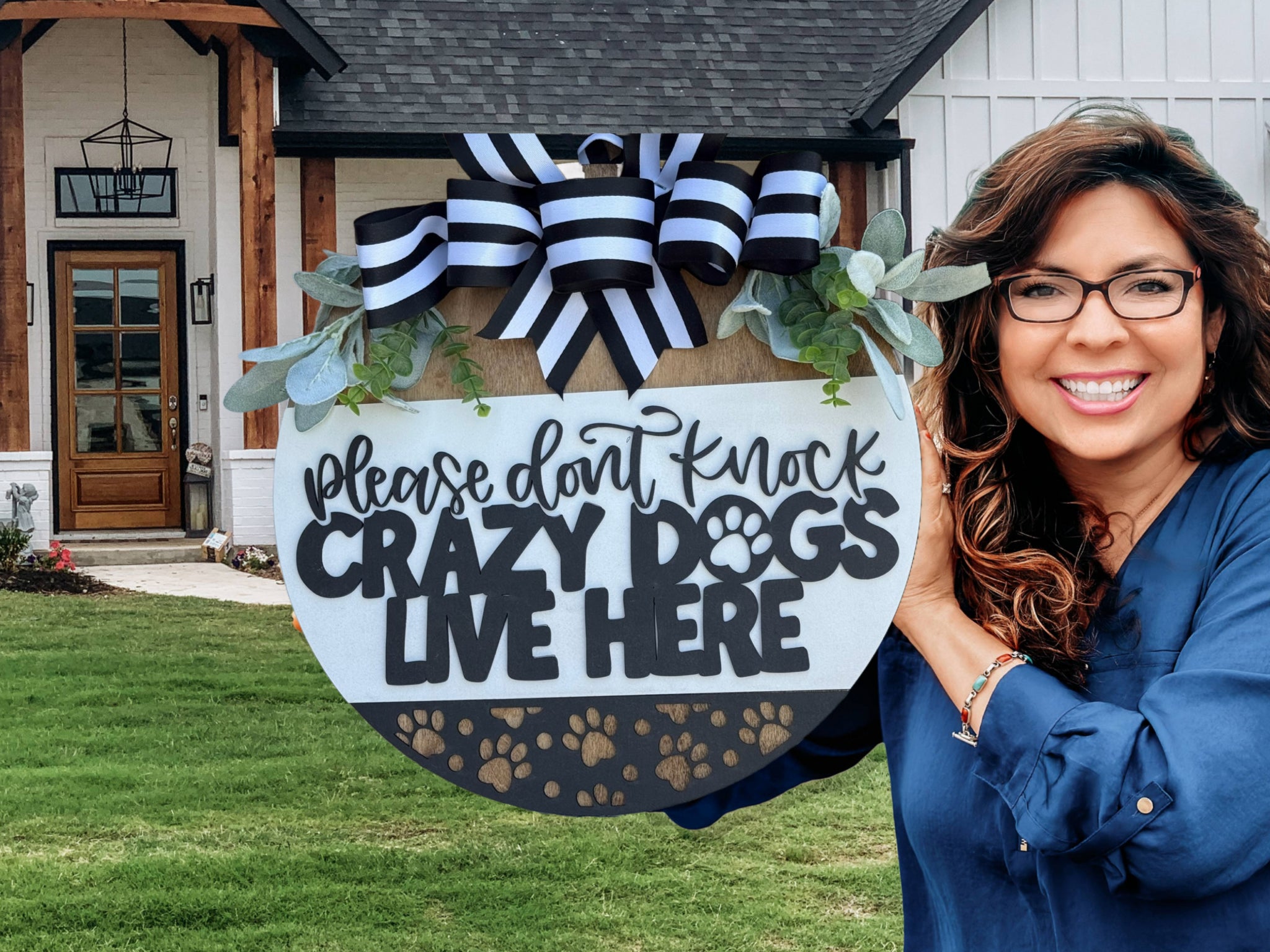 Smiling woman with long brown hair holds a decorative round sign that reads, Please dont knock, crazy dogs live here, in front of a modern house with green grass and a porch.