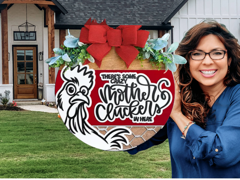A smiling woman with long brown hair and glasses holds a round sign with a chicken graphic, a red bow, and the text Theres some crazy mother cluckers in here, standing in front of a modern house.