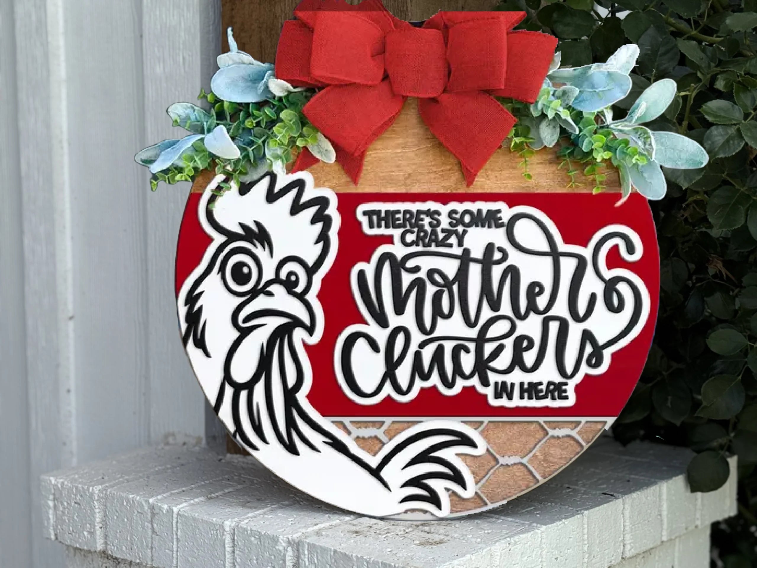 A decorative sign with a rooster illustration, greenery, and a red bow reads Theres some crazy mother cluckers in here. The background includes red, brown, and white patterns, and the sign is propped against an outdoor wall.