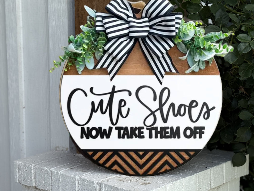 A round wooden sign decorated with a black and white striped bow and greenery reads, Cute Shoes NOW TAKE THEM OFF. The sign sits on a white brick surface near a wall.