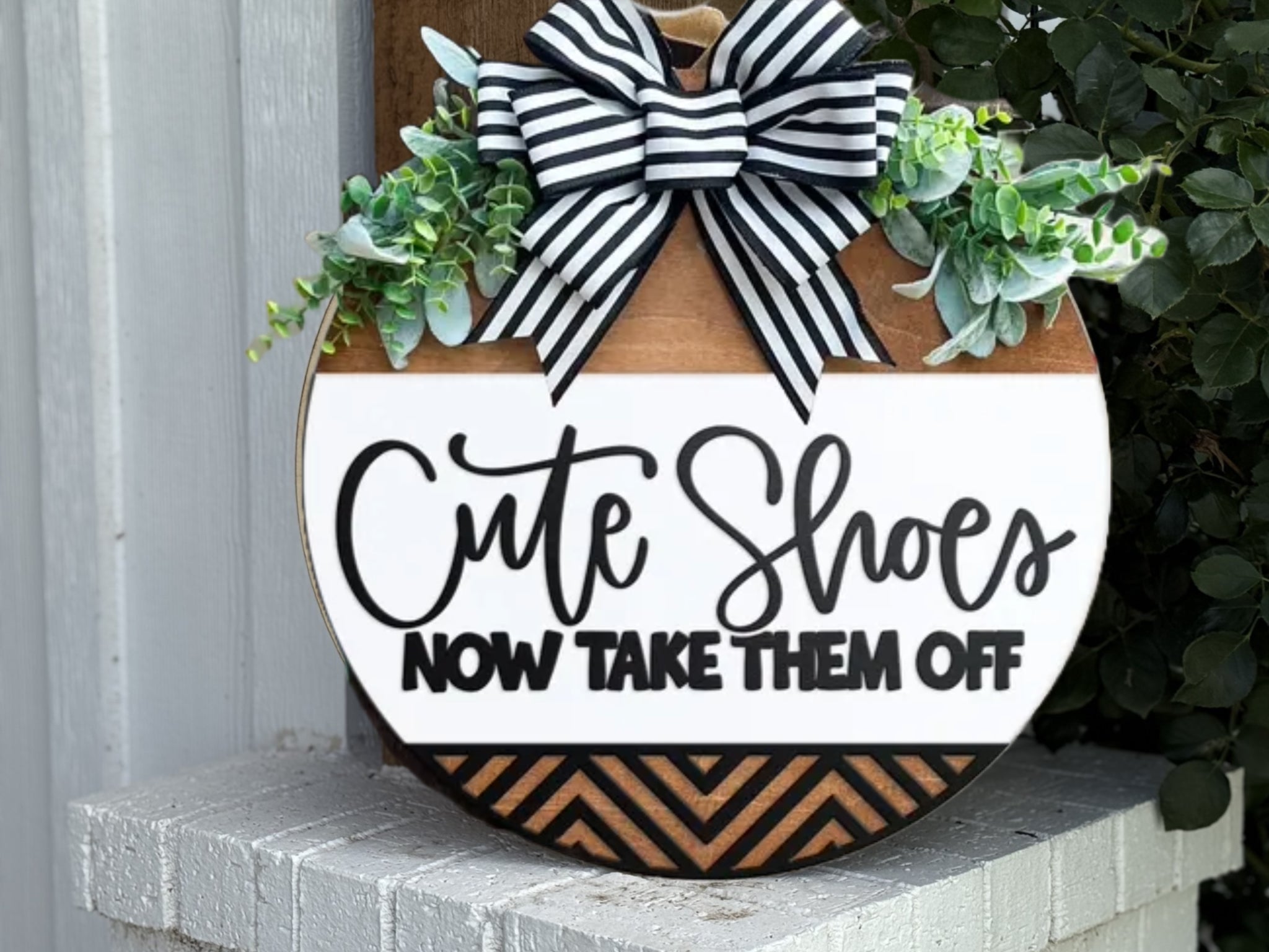 A round wooden sign decorated with a black and white striped bow and greenery reads, Cute Shoes NOW TAKE THEM OFF. The sign sits on a white brick surface near a wall.