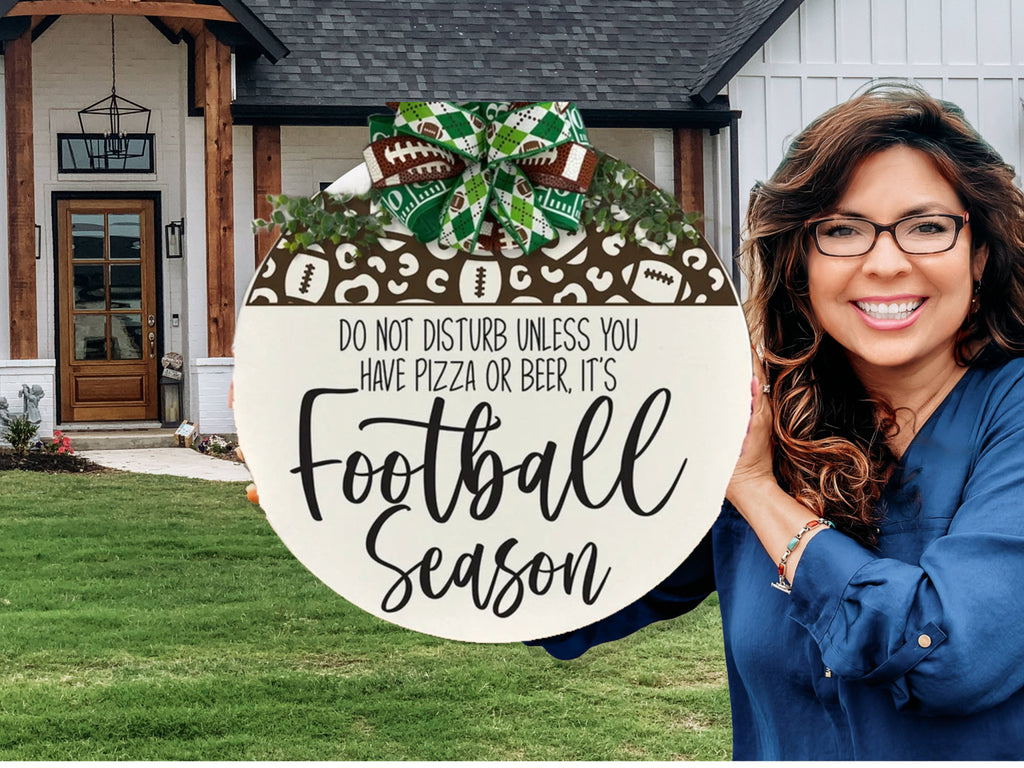 A smiling woman with long brown hair and glasses holds a round sign decorated with football motifs and a bow. The sign reads, “Do not disturb unless you have pizza or beer, it’s Football Season.” A house with a porch is in the background.