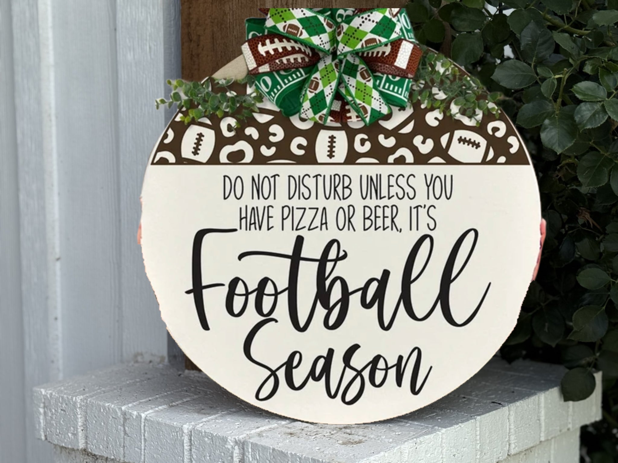 A round door sign with a bow and football-themed decorations says, Do not disturb unless you have pizza or beer, it’s Football Season. The sign is displayed on a white brick surface near some green leaves.