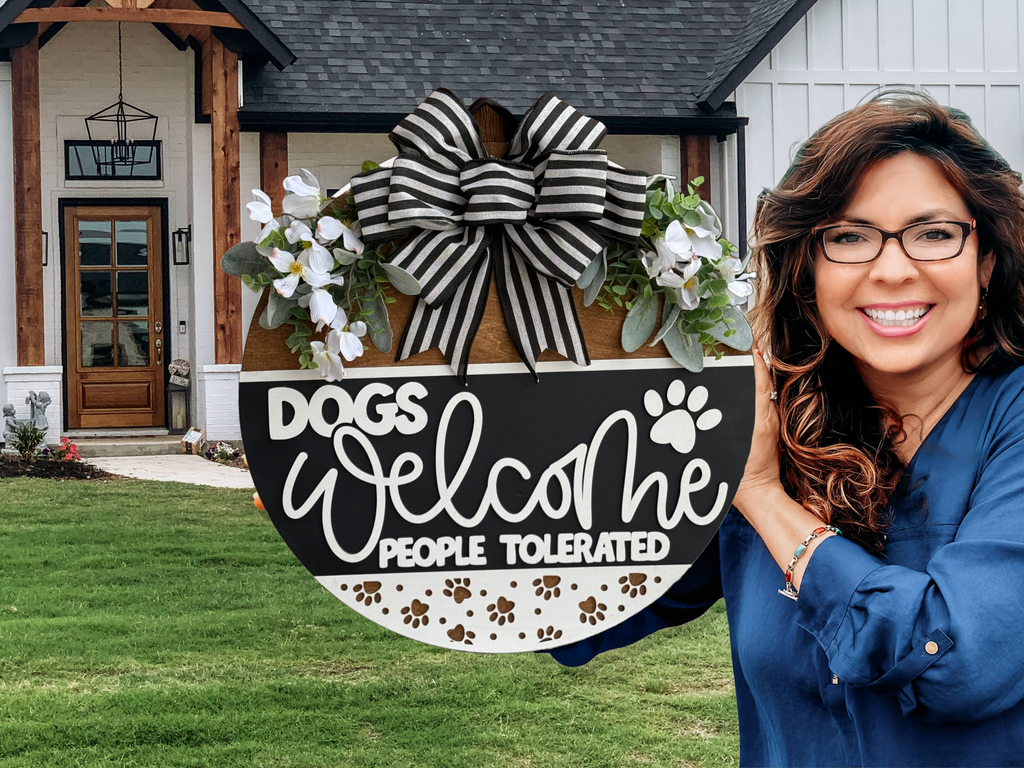 A smiling woman holds a round sign decorated with flowers and a striped bow that reads, Dogs Welcome, People Tolerated. She stands in front of a modern house with a green lawn.