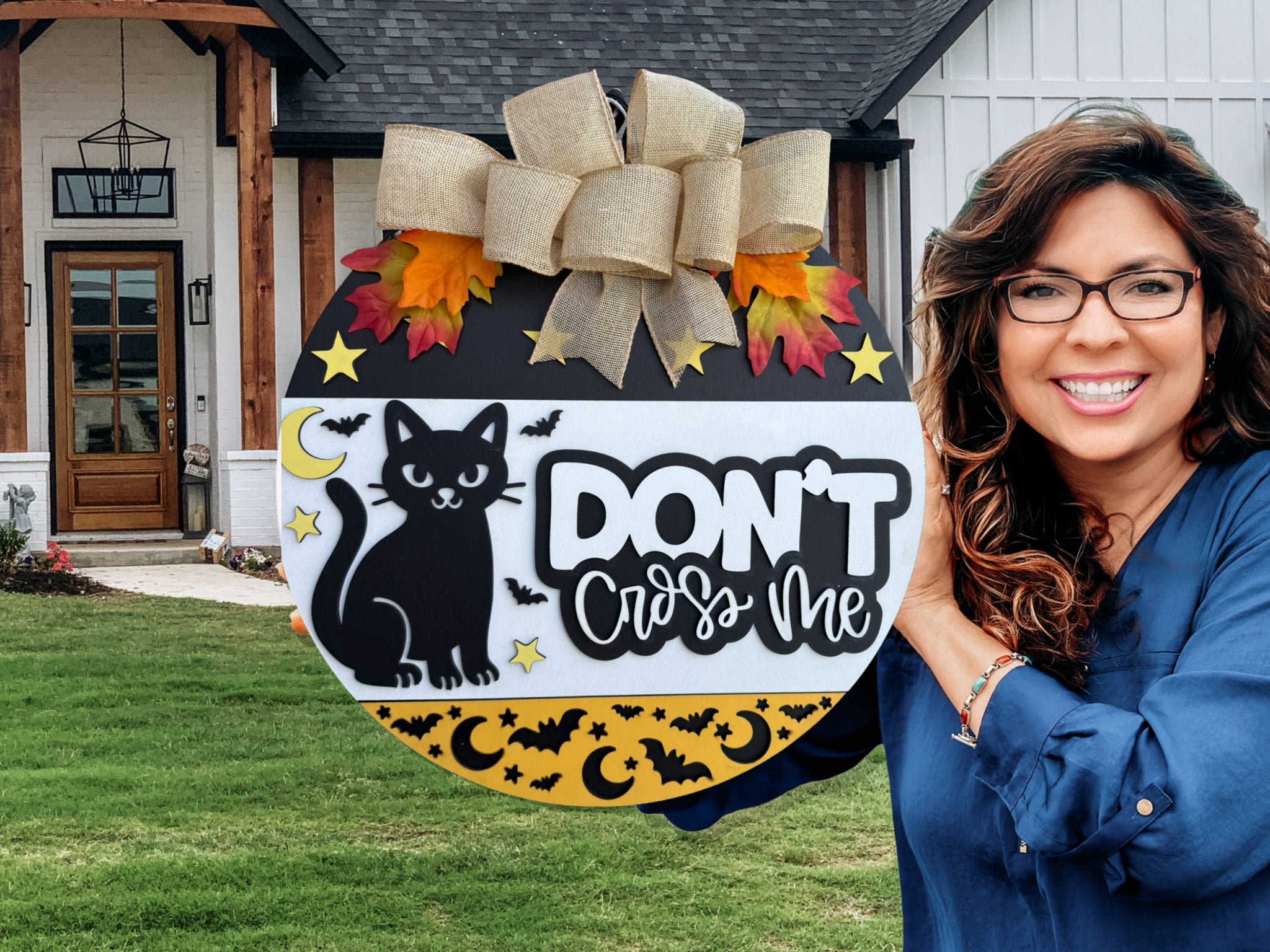 A smiling woman with wavy brown hair and glasses holds a round Halloween sign decorated with a black cat, stars, moons, bats, and the words DONT cross me in front of a modern house.