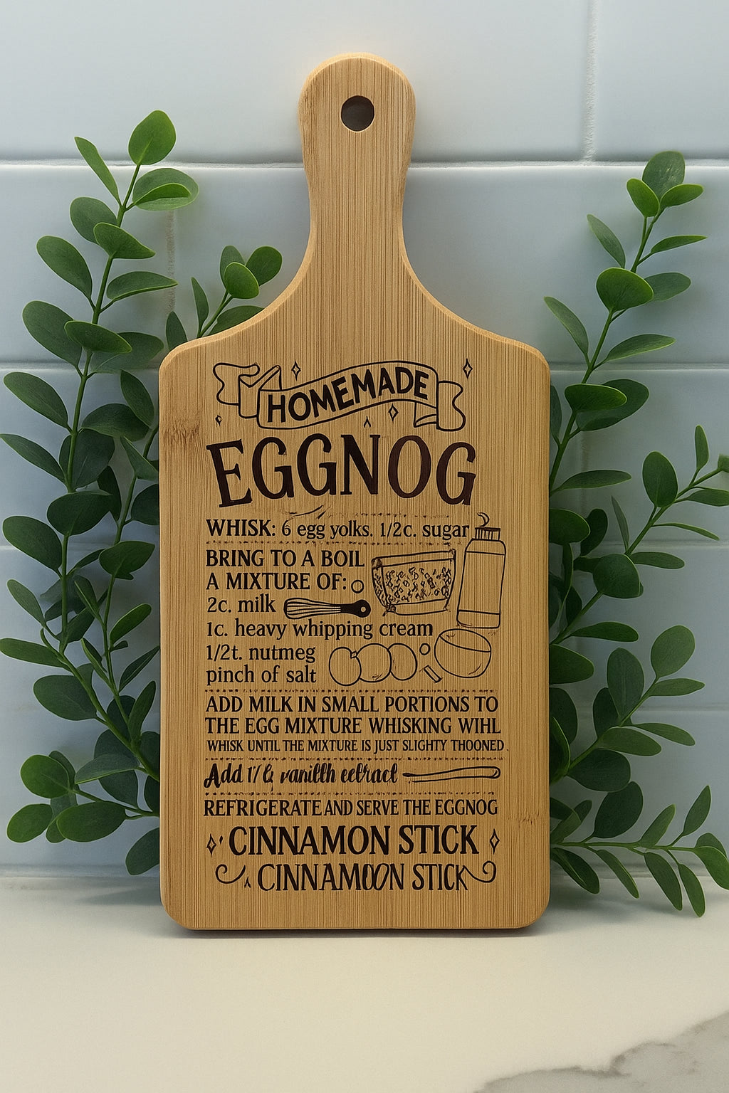 A wooden cutting board with an engraved homemade eggnog recipe, illustrated with a mixing bowl, whisk, and measurements, stands upright against a tiled wall with green leafy plants on either side.