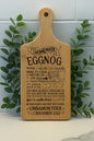 A wooden cutting board with an engraved homemade eggnog recipe, illustrated with a mixing bowl, whisk, and measurements, stands upright against a tiled wall with green leafy plants on either side.