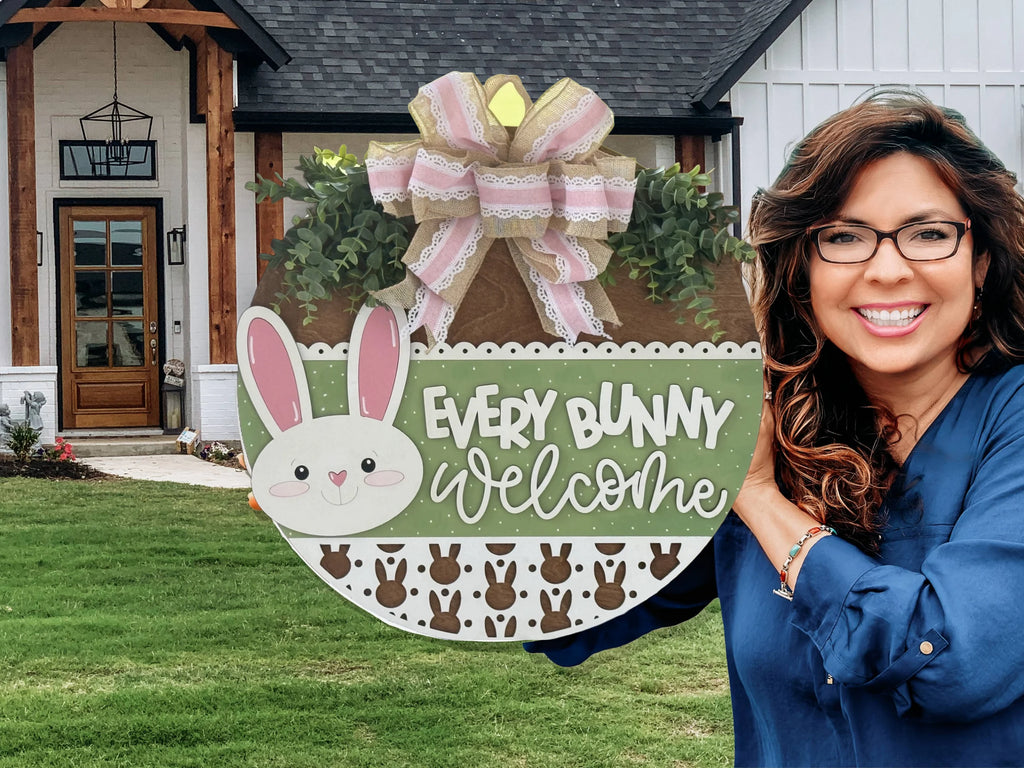 A smiling woman with wavy brown hair and glasses holds a round Easter-themed sign reading “Every Bunny Welcome,” decorated with a bunny face, green and white patterns, and a large pastel ribbon. She stands in front of a modern house with a wooden door.