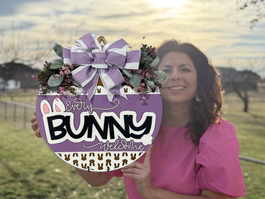 A woman in a pink shirt holds a round sign with a purple background featuring the phrase Every Bunny Welcome decorated with small bunny shapes. The top of the sign has a large bow with lavender ribbons and floral accents. The background shows a grassy field and a cloudy sky.