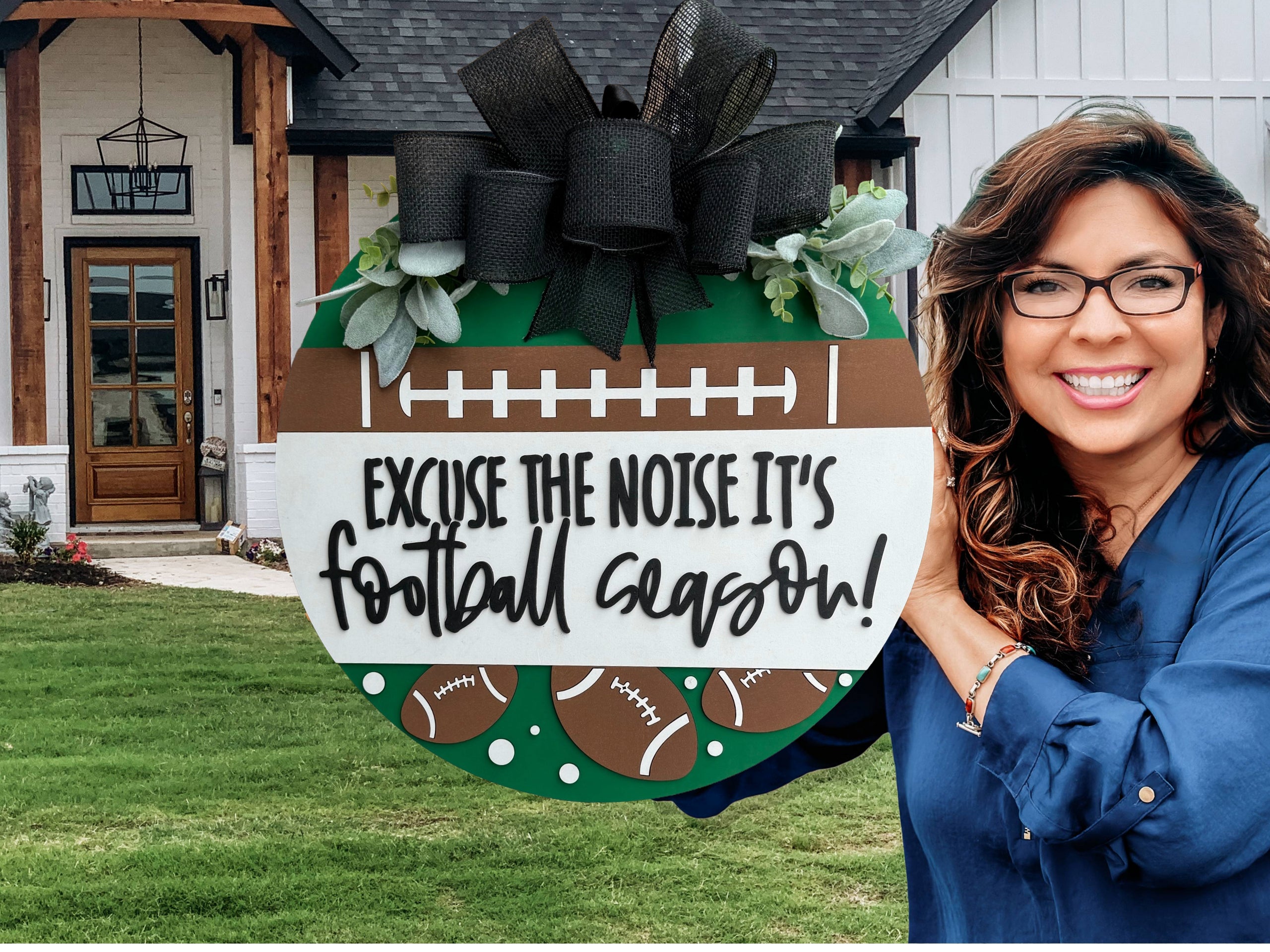 A round sign decorated with faux greenery and footballs reads, “Excuse the noise it’s football season” in bold, playful lettering. The sign features a football field design and is displayed against a white textured surface.